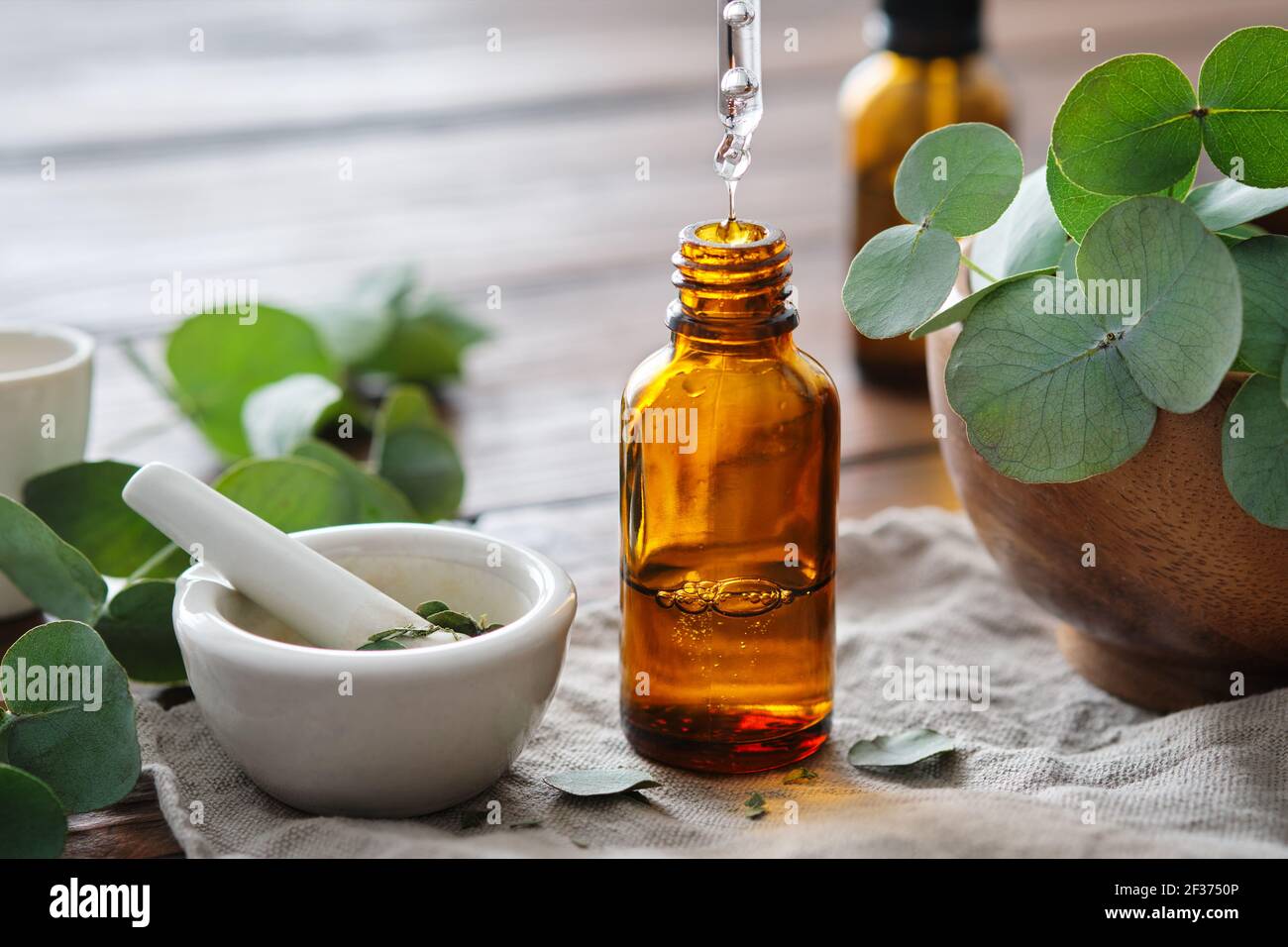 Flasche Eukalyptusöl, Mörser und Holzschale mit grünen Eukalyptusblättern. Nahaufnahme eines Tropfen ätherischen Eukalyptus-Öls, der aus einem Glastropfen tropft Stockfoto