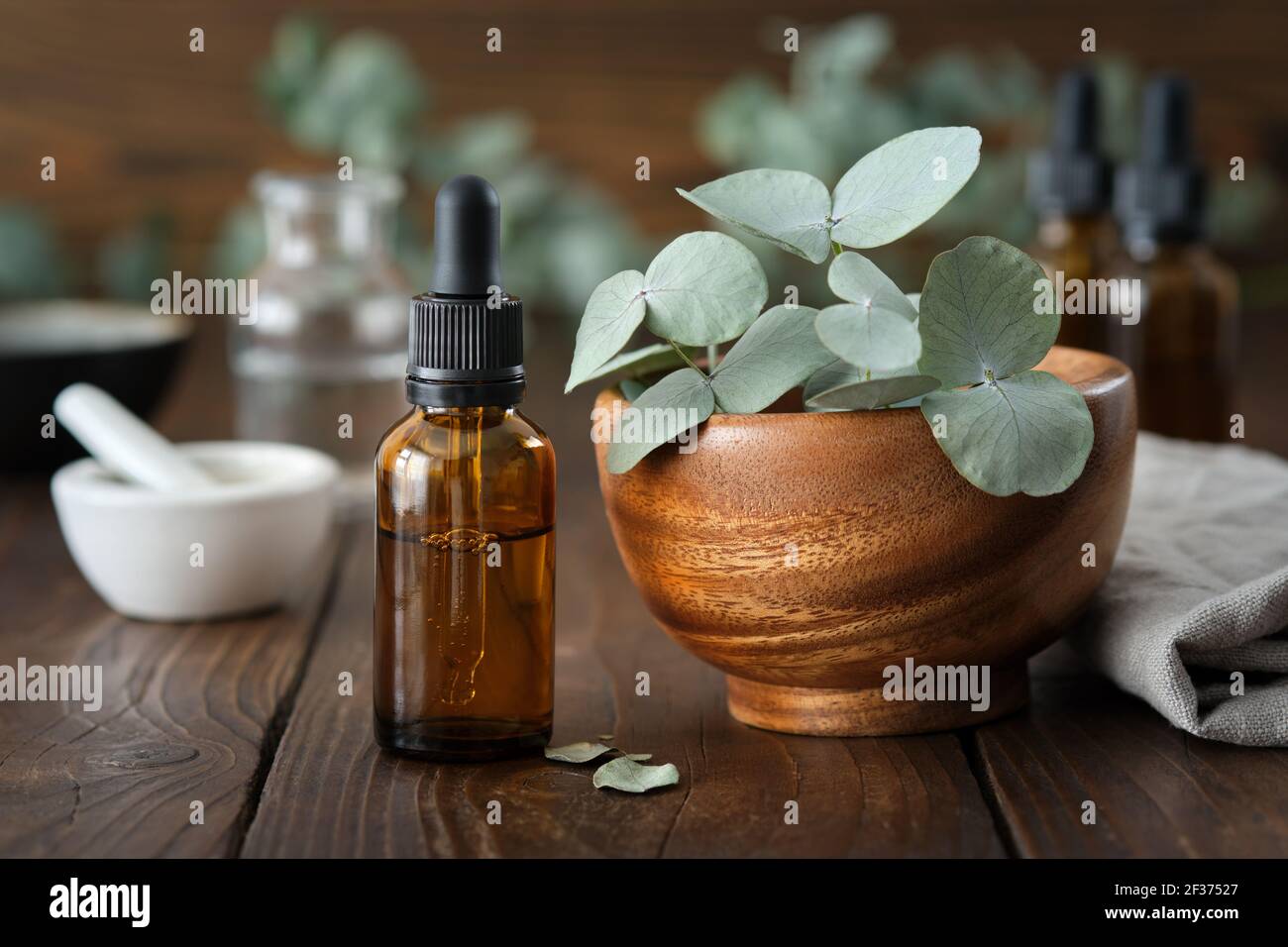 Tropfflasche mit ätherischem Eukalyptus-Öl und Holzschüssel mit grünen Eukalyptus-Blättern. Mörtel und Ölflaschen auf dem Hintergrund, nicht im Fokus. Stockfoto