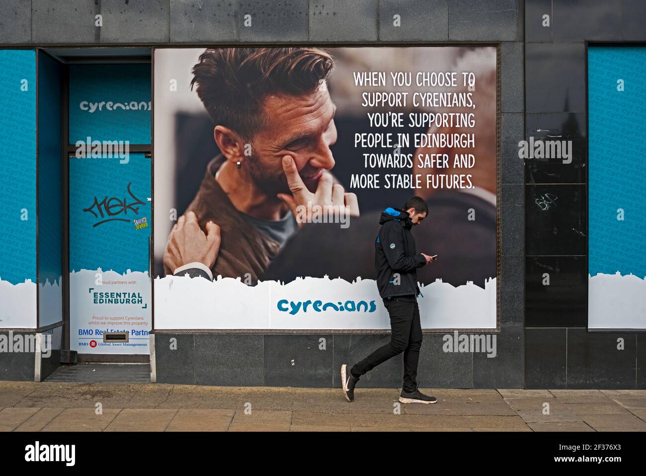 Junger Mann, der sein Telefon anschaut, während er an einer Werbung für die Cyrenians in einem leeren Laden in der Princes Street, Edinburgh, Schottland, Großbritannien, vorbei geht. Stockfoto