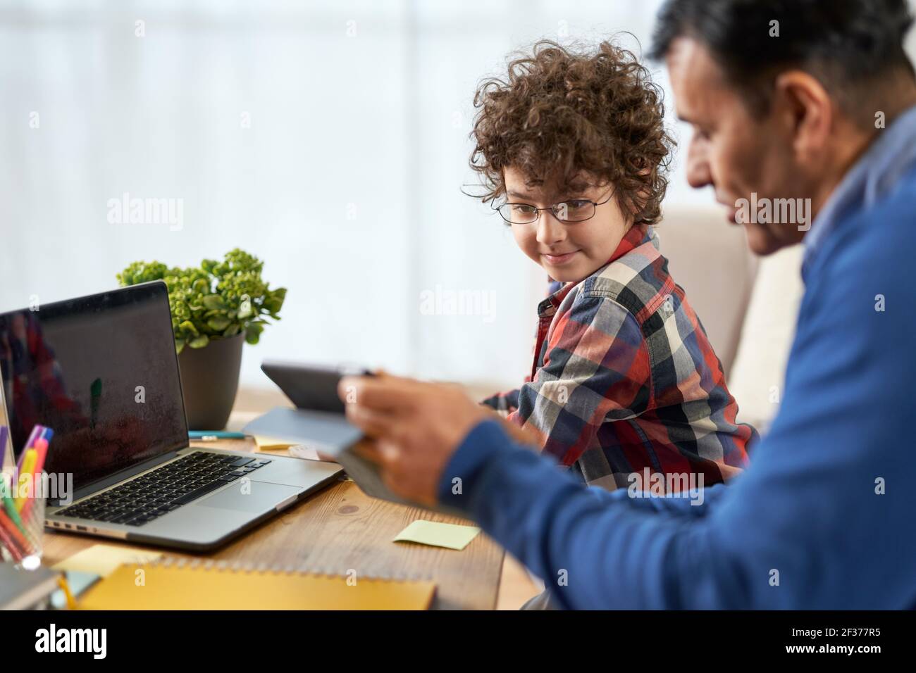 Lehrkind. Porträt des niedlichen kleinen hispanischen Schuljungen, der zusammen mit seinem Vater Hausaufgaben macht und Technologie verwendet, während er zu Hause am Schreibtisch sitzt Stockfoto