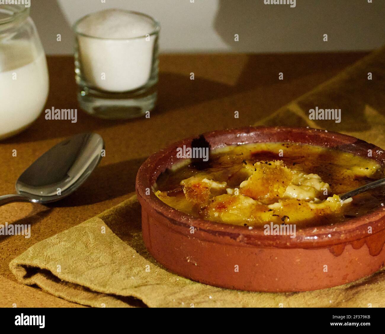 Löffel in katalanische Sahne getaucht auf einer Serviette mit einer Tasse Zucker und einer Tasse Milch dekoriert. Auf dem Holztisch Stockfoto