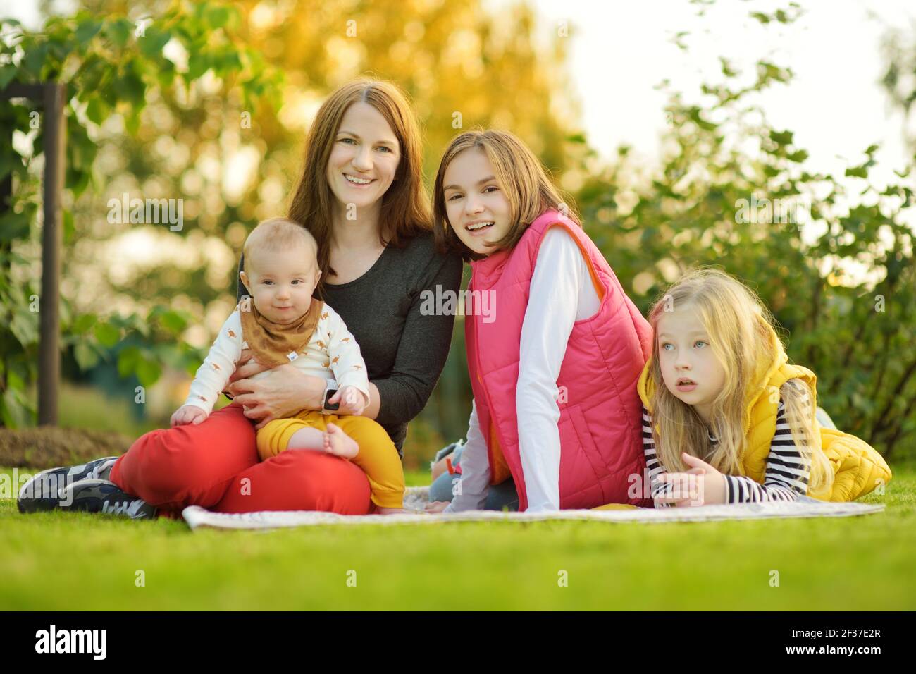 Mutter und drei Kinder haben Spaß am Herbsttag im Stadtpark. Liebenswert Baby Junge wird von seiner Mama gehalten. Zwei ältere Schwestern umarmen ihre Mutter und BA Stockfoto