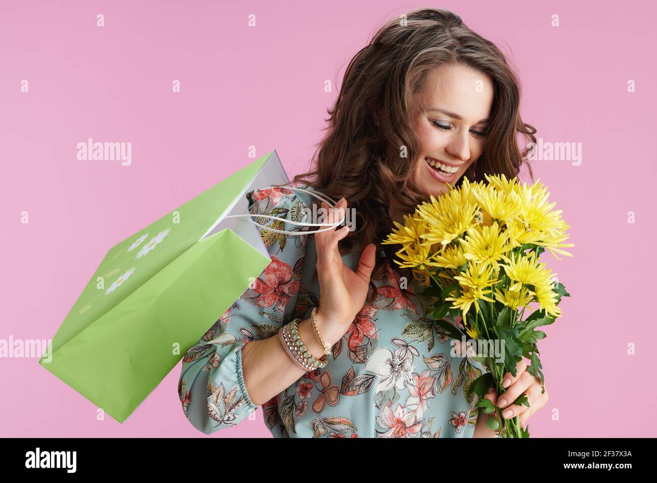 Lächelnd stilvolle Frau im Blumenkleid mit gelben Chrysanthemen Blumen und grüne Einkaufstasche isoliert auf rosa. Stockfoto