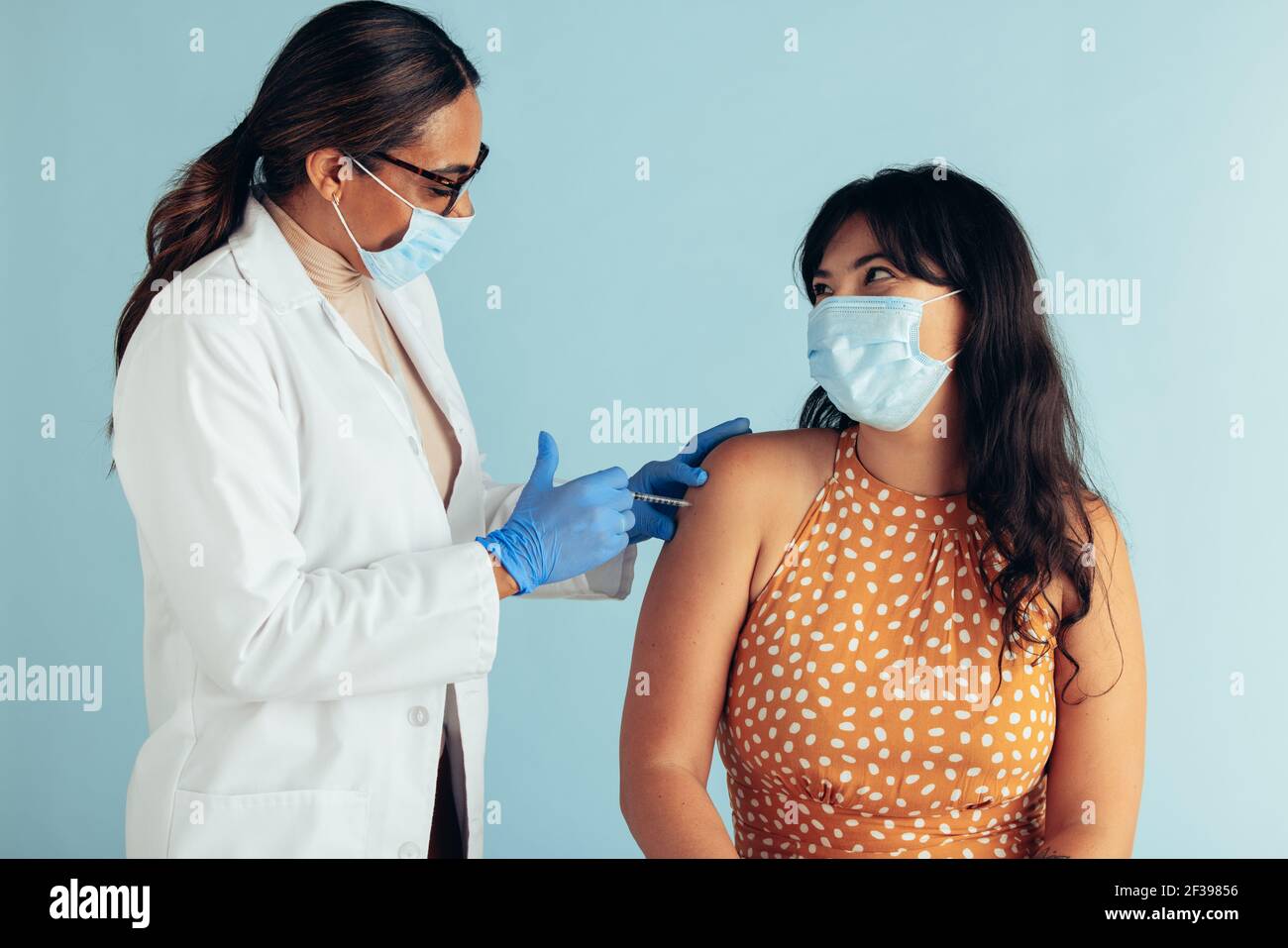 Ärztin und Patientin tragen Gesichtsmasken während der Impfung. Arzt, der Frauen vor blauem Hintergrund einen Impfstoff gibt. Stockfoto