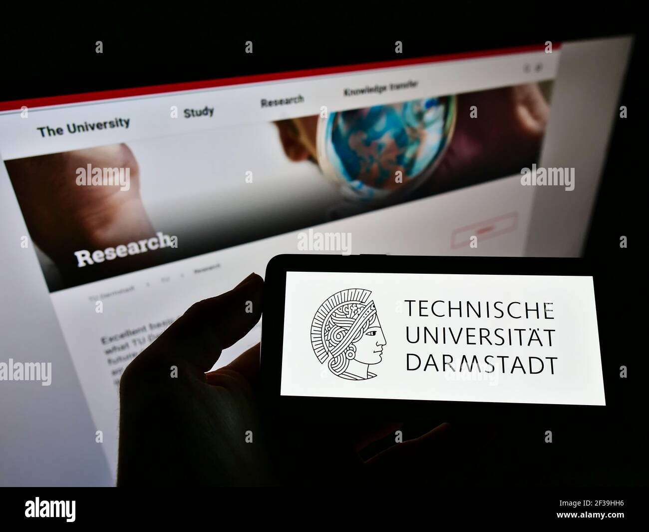 Person, die das Mobiltelefon mit dem Logo der Technischen Universität Darmstadt auf dem Bildschirm vor der Webseite hält. Konzentrieren Sie sich auf die Anzeige des Mobiltelefons. Stockfoto