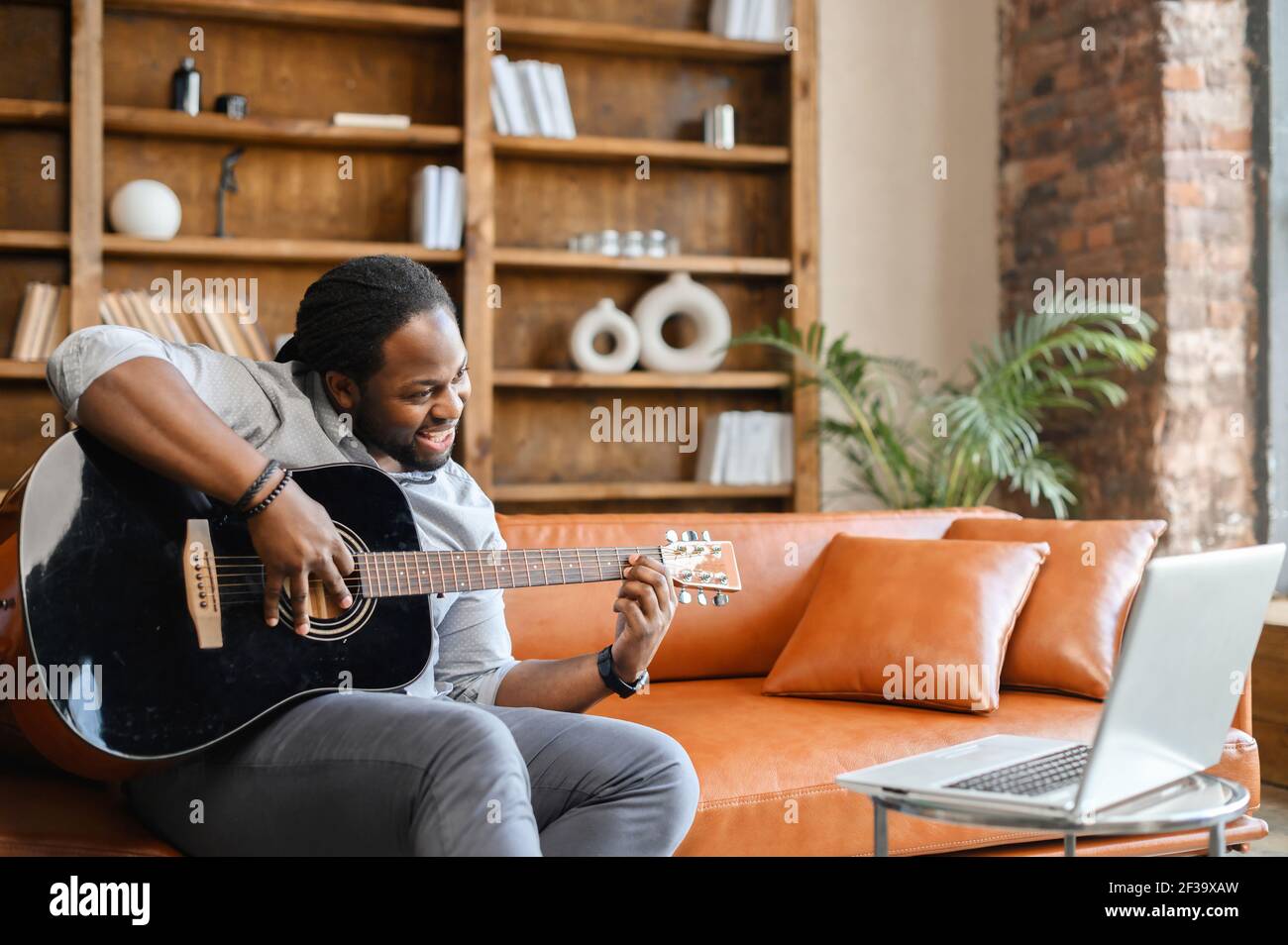 Afrikanischer Mann sitzt auf dem Ledersofa mit Gitarre in einem kreativen Raum oder zu Hause, spielt ein Lied für seine Freundin, lehrt online, zeigt Meisterkurs, Aufnahme Song, lernt Song aus einem Video Stockfoto