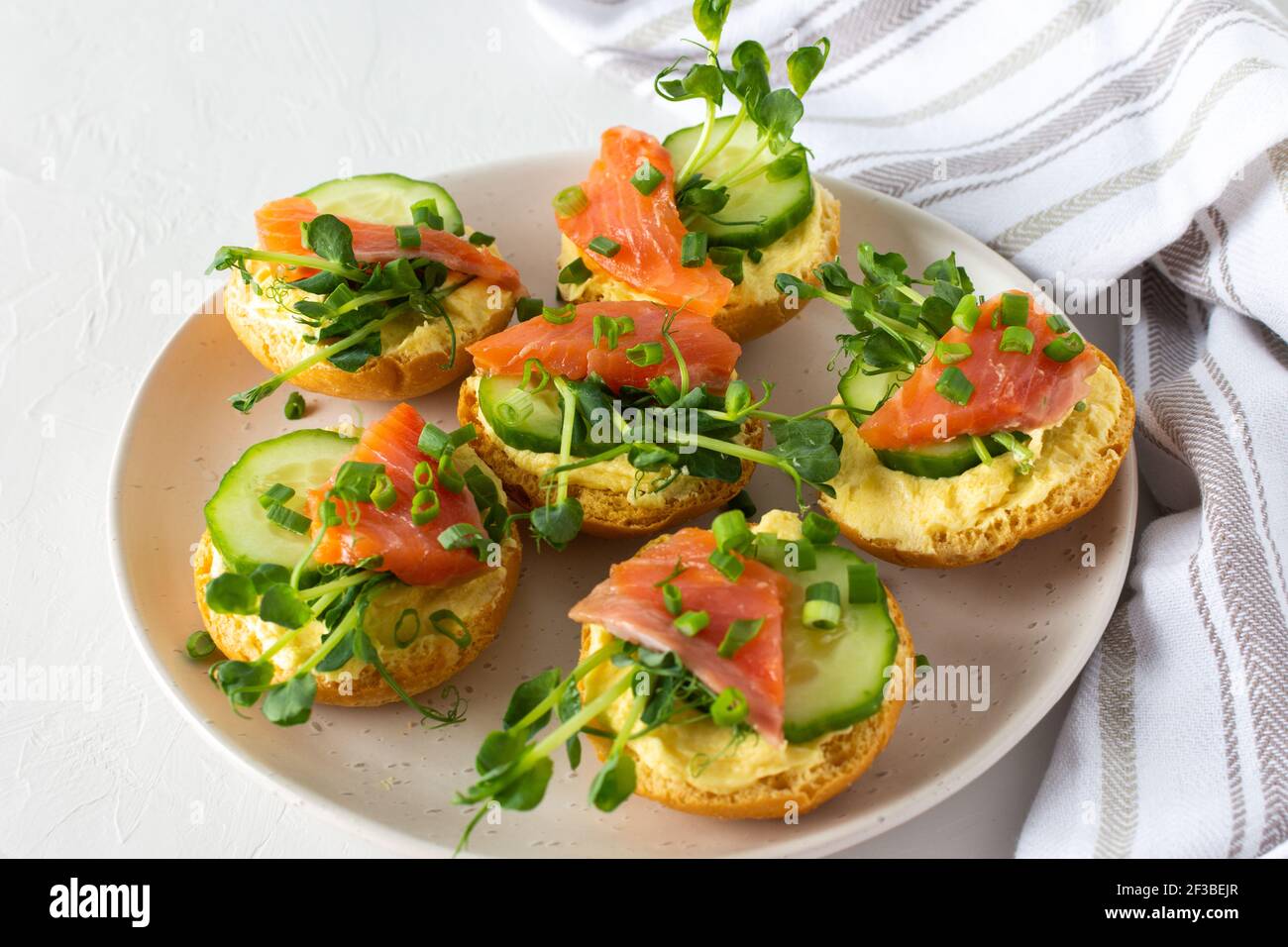 Leckere herzhafte Vorspeise, Lachsbraten auf einem weißen Teller, rundes gebratenes Brötchen mit rotem Fisch, Gurke, Sauce Hollandaise und Erbsensprossen Stockfoto