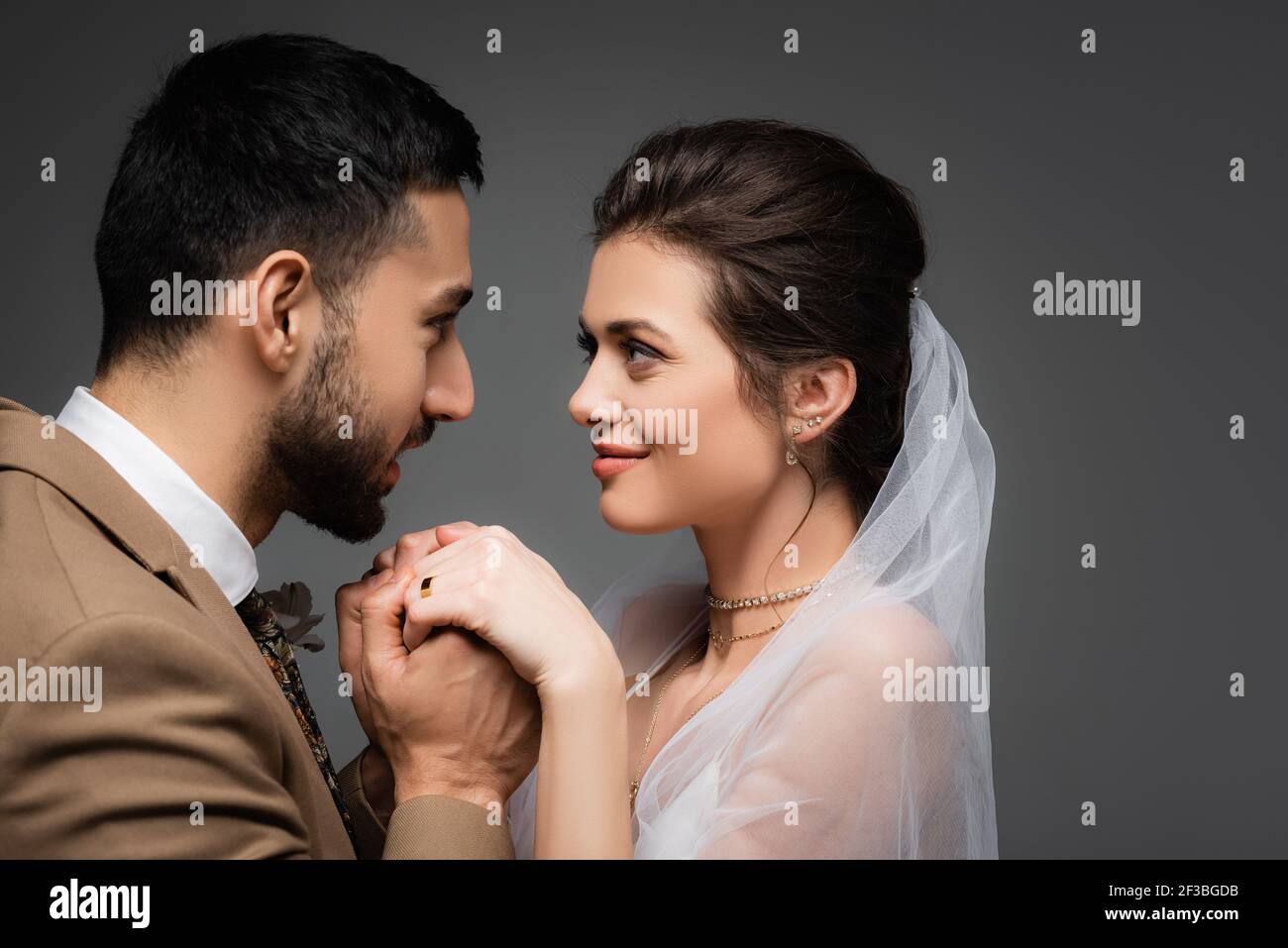 Eleganter muslimischer Mann, der die Hände einer glücklichen Braut isoliert hält Grau Stockfoto