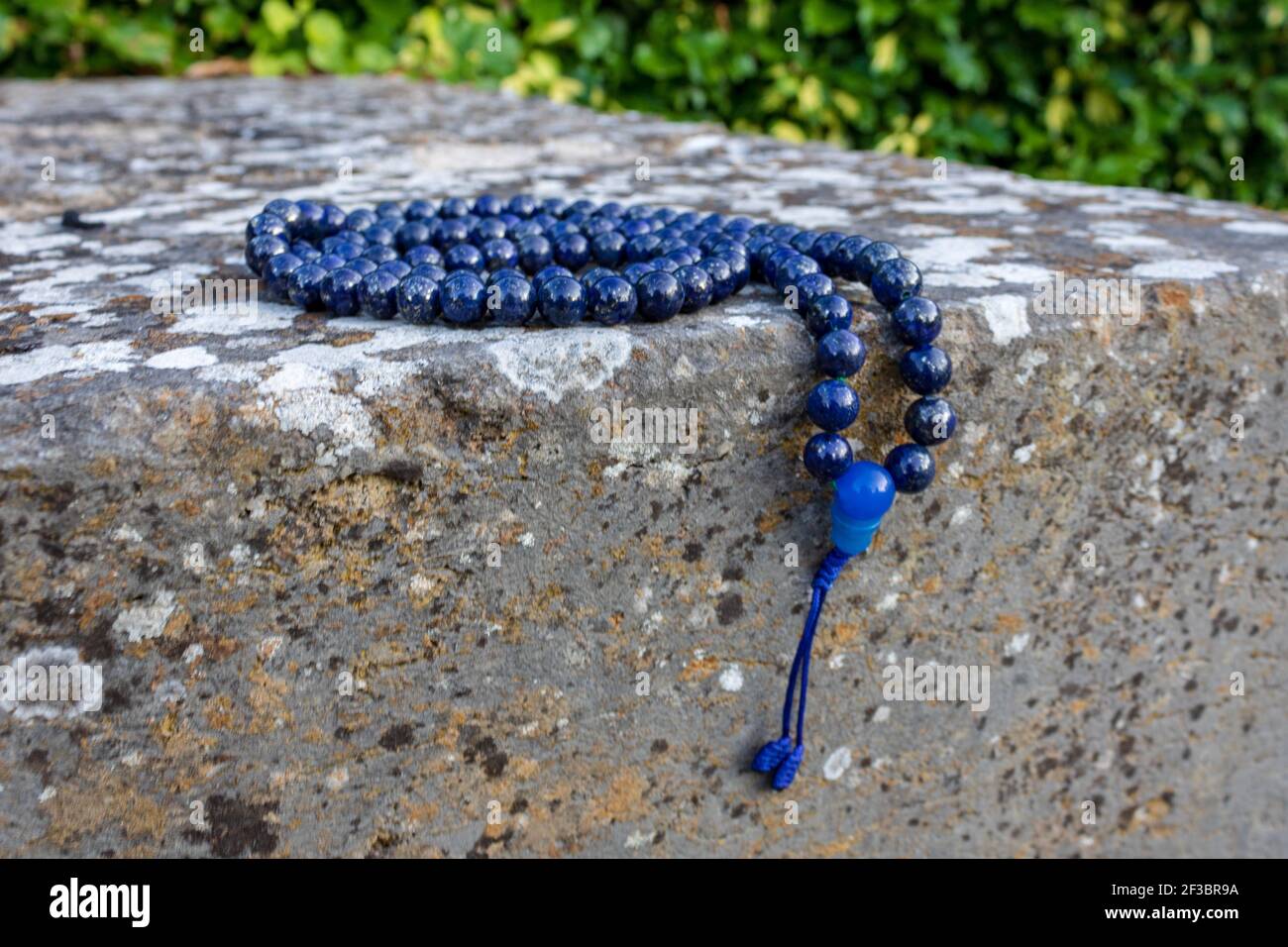 Nahaufnahme von blauen mala Perlen (Lapislazuli) auf Stein mit grünen Blättern im Hintergrund. Achtsamkeit und Meditationszubehör. Stockfoto