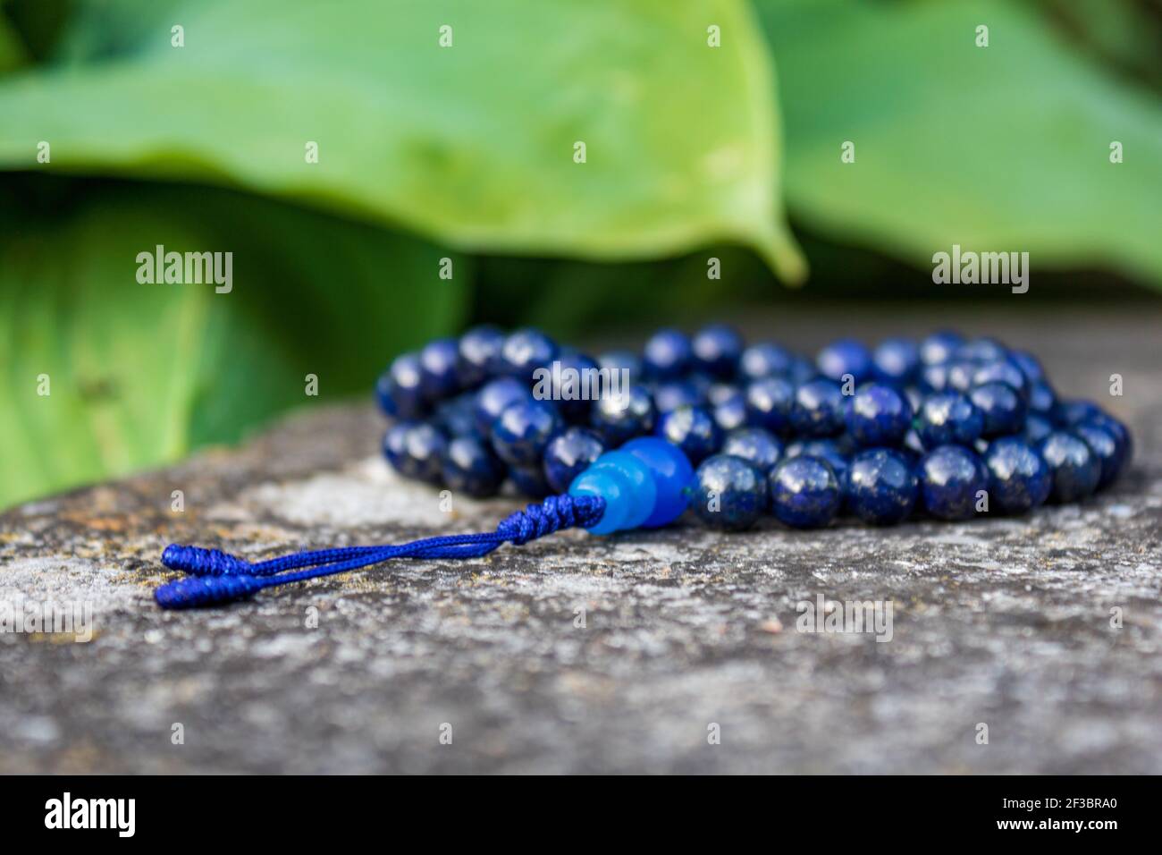 Blaue mala Perlen (Lapislazuli) auf Stein mit grünen Blättern im Hintergrund. Yoga und Japa Meditation Zubehör. Stockfoto