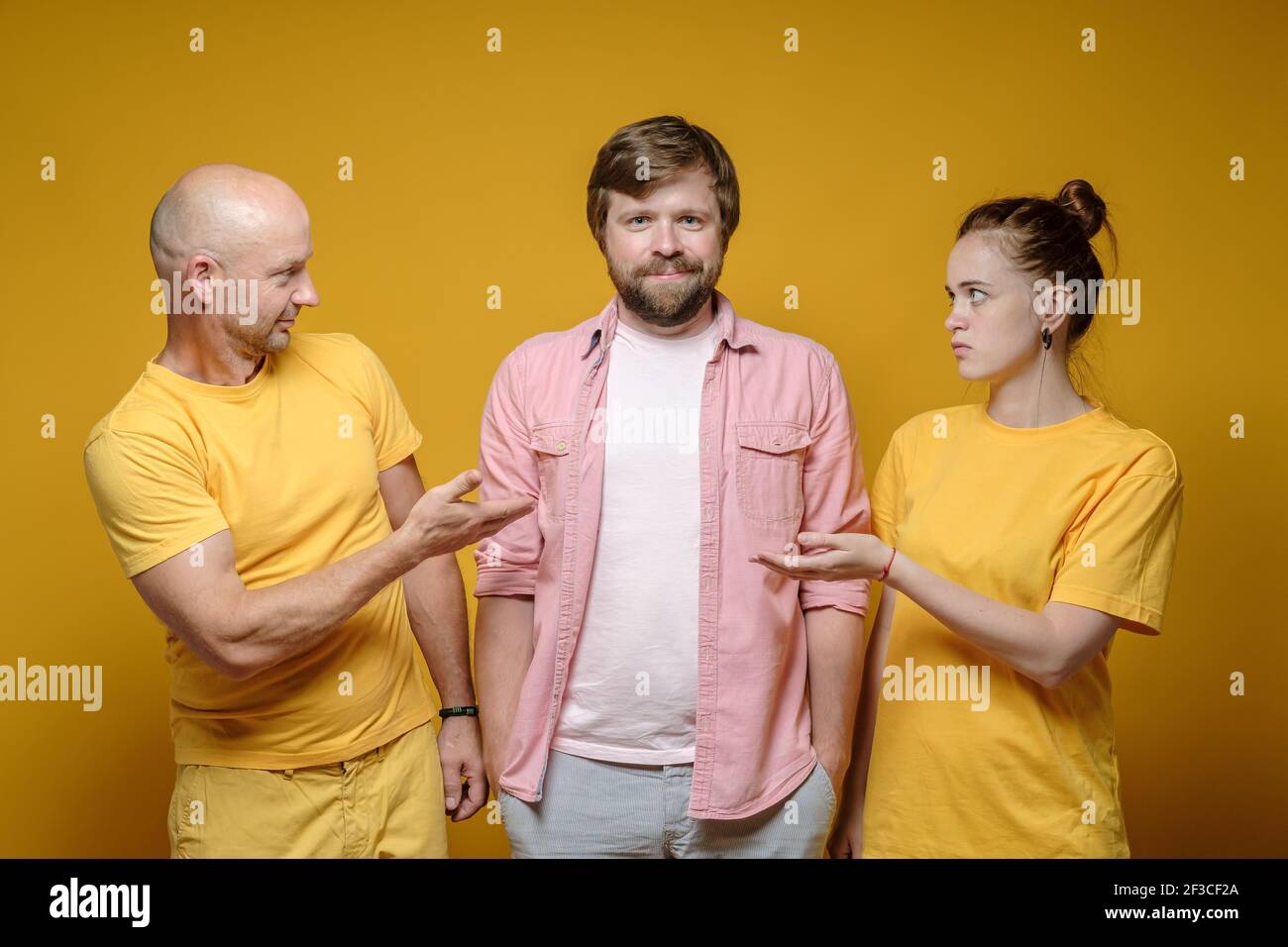 Mann und eine Frau sehen ihren zufriedenen und lächelnden Freund in Verwirrung an. Gelber Hintergrund. Stockfoto