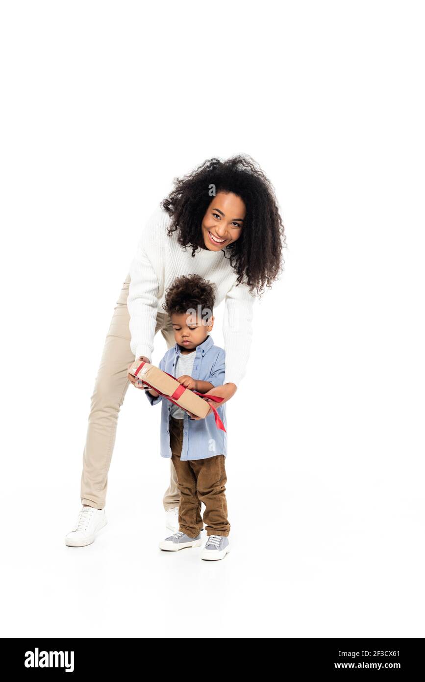 Fröhliche afroamerikanische Frau, die Kamera in der Nähe von Sohn mit Geschenkbox auf weiß Stockfoto