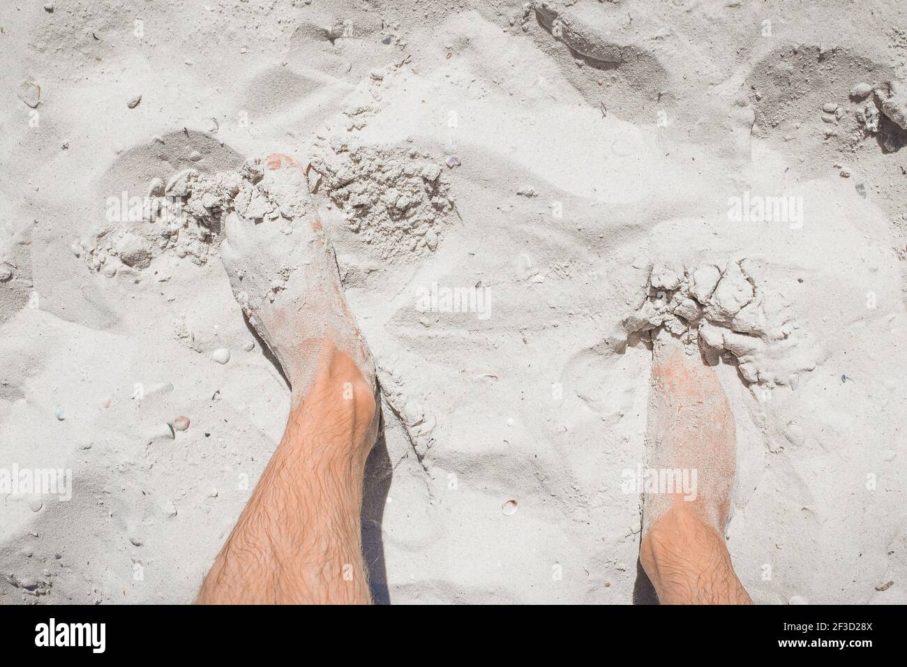 Die Beine des jungen Mannes stehen entspannt auf dem weißen Strandsand, der Blick von oben. Stockfoto