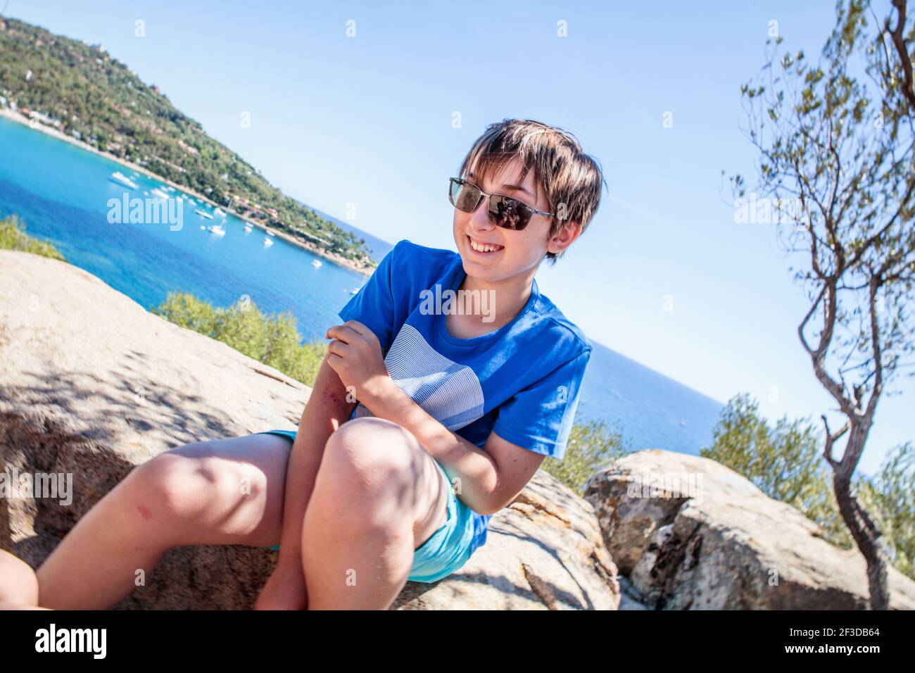 Ein 10-jähriger Junge sitzt auf einigen Felsen an der Küste im Urlaub auf Sardinien. Blick auf das Meer und die Berge Stockfoto