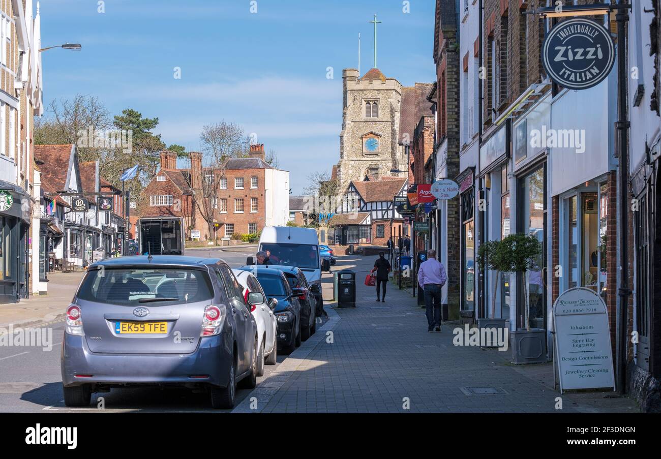 Pinner Village High Street mit Geschäften, St. John the Baptist Pfarrkirche, geparkten Autos und ein paar Menschen zu Fuß auf dem Bürgersteig. NW London, England Stockfoto