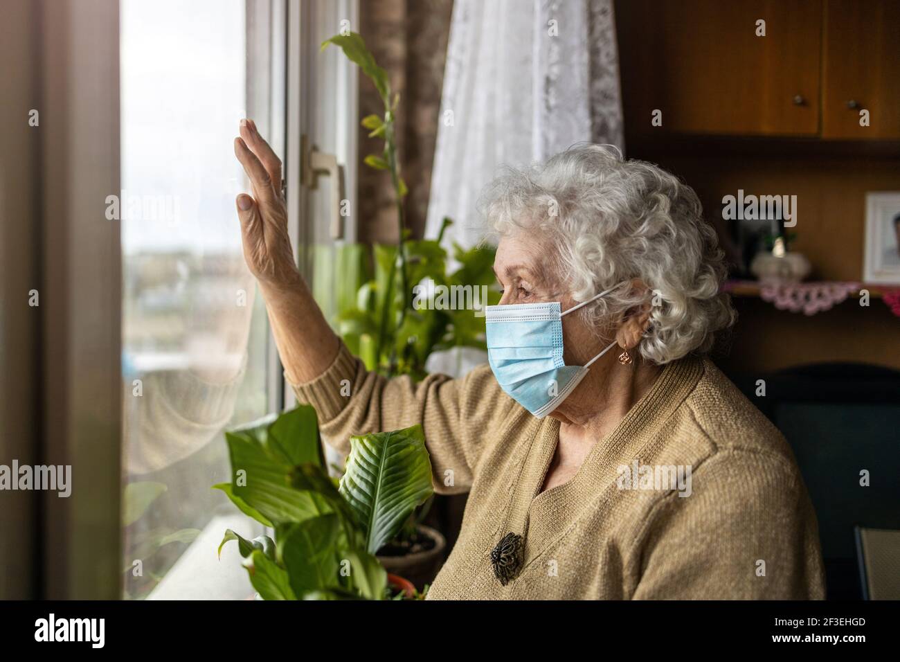 Ältere Frau mit Gesichtsmaske, die aus dem Fenster schaut Zu Hause Stockfoto