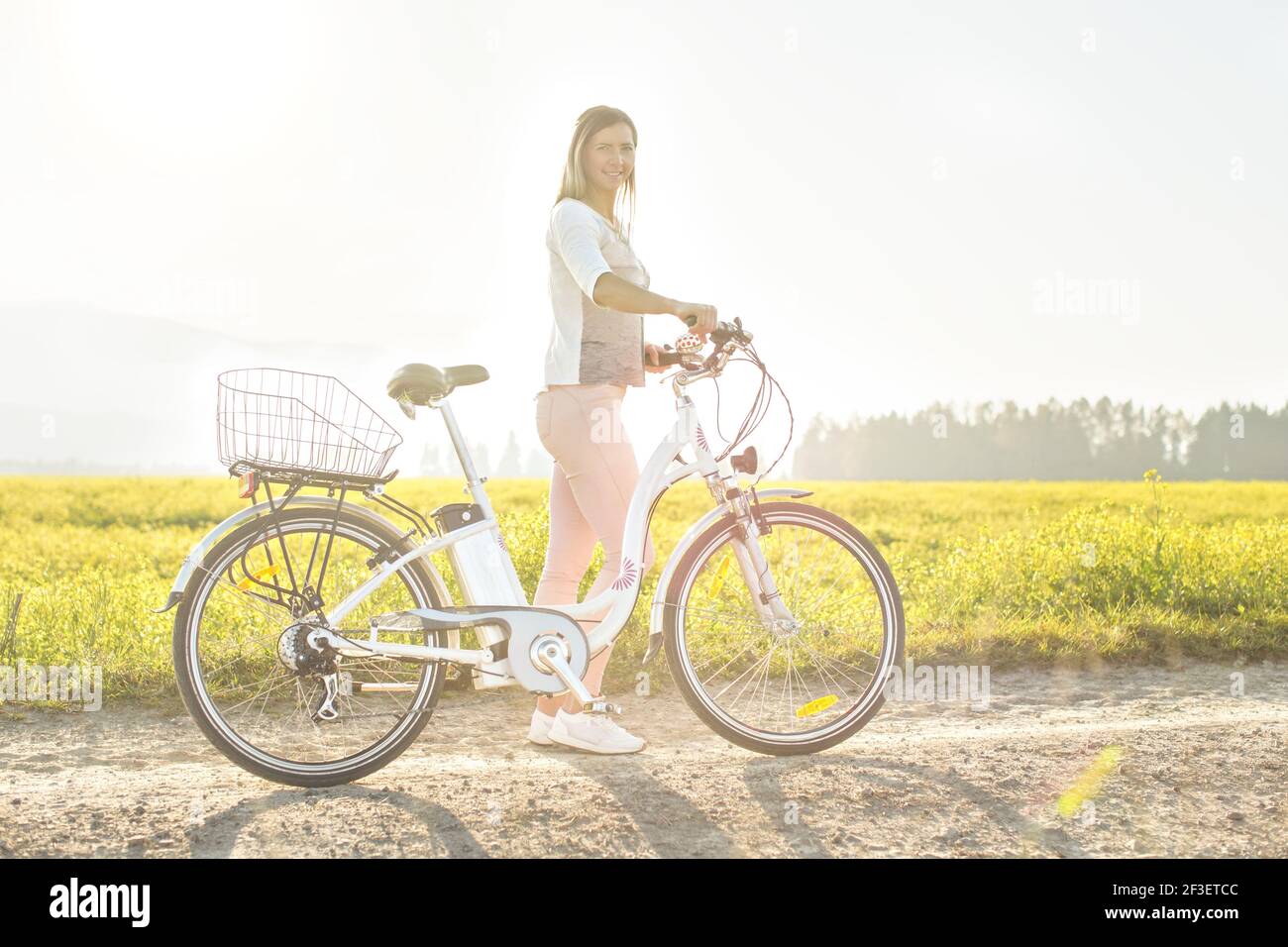 Junge Frau neben ihrem Elektro-Fahrrad über staubige Landstraße stehen, starke Nachmittag Sonne Hintergrundbeleuchtung im Hintergrund leuchtet auf gelben Blumen Feld, V Stockfoto