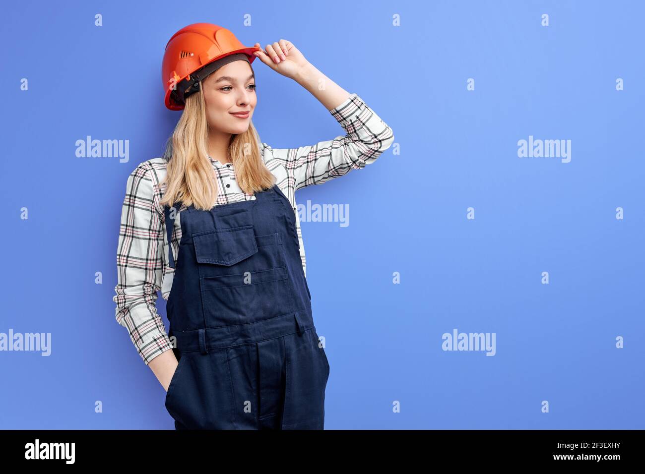 Architektin Frau in Overalls isoliert über blauem Studiohintergrund, junge Frau in orangefarbenem Hut posiert vor der Kamera und schaut zur Seite, arbeitet als su Stockfoto