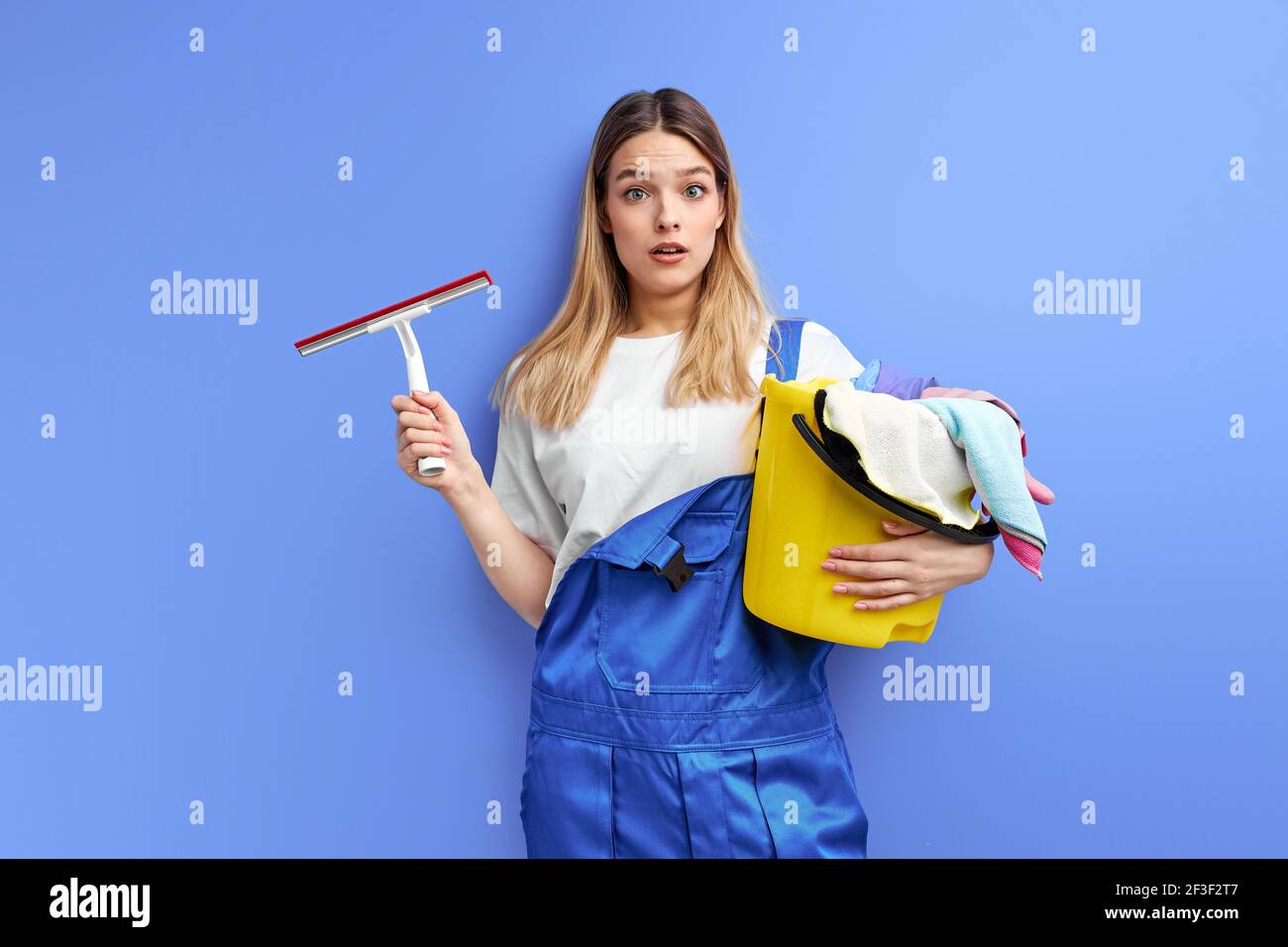 Frau hält Eimer mit Gruppe von Reinigungsmitteln. Junge Dame in Overalls stehen in Schock durch Schmutz, Haushaltsgeräte, Frühjahrsputz, Aufräumen Stockfoto