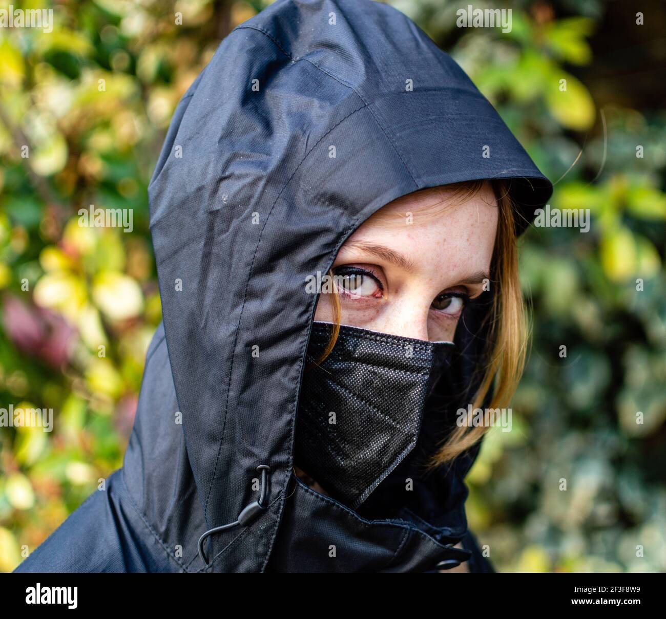 Nahaufnahme eines blonden Mädchens, mit schwarzer Gesichtsmaske und Kapuze, verschwommener Hintergrund Stockfoto
