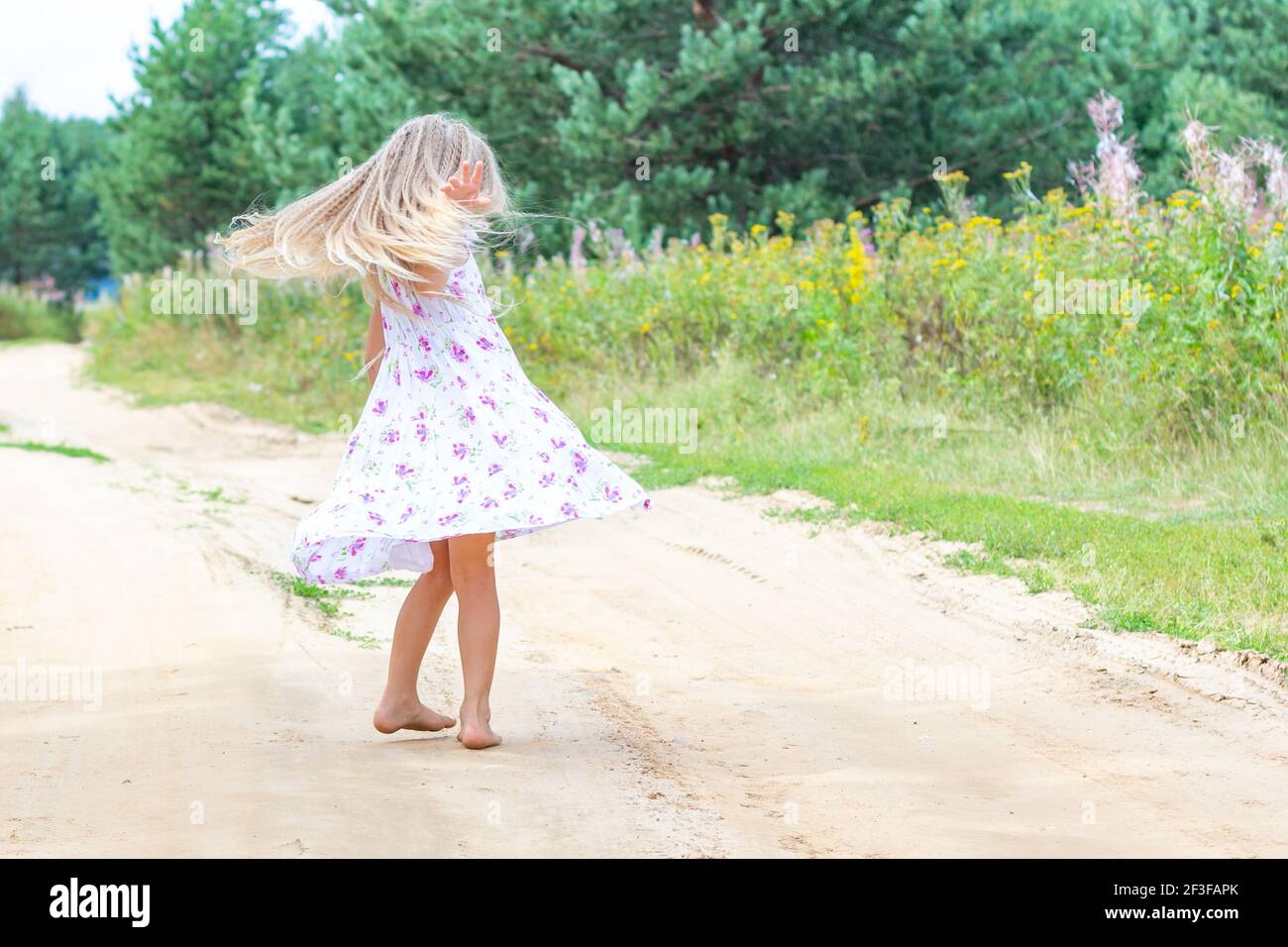 Ein Mädchen mit langen blonden Lockenhaaren tanzt und dreht sich auf einer Waldstraße. Stockfoto