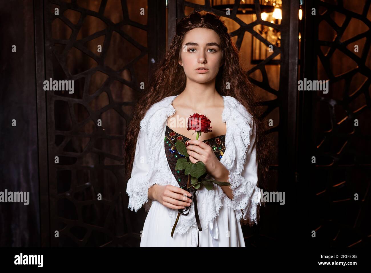 Eine junge schöne Brünette mit langen lockeren Haaren in einem Weißer Peignoir in der Art und Weise des 19th. Jahrhunderts hält Eine scharlachrote Rose in ihren Händen - ein Cosplay für Das Broadway Musical das Phantom der Oper Stockfoto