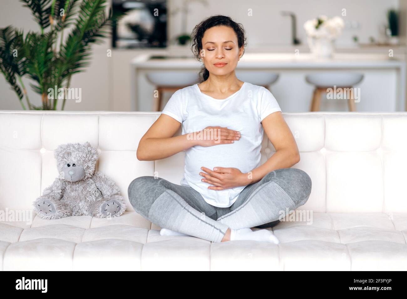 Erstaunliche gemischte Rasse Erwachsene schwangere Frau trägt weißes T-Shirt und Leggings entspannt im Wohnzimmer auf dem Sofa zu Hause, meditiert in der Lotus-Position, sanft umarmt ihren Bauch, denken Sie an Baby, Augen geschlossen Stockfoto