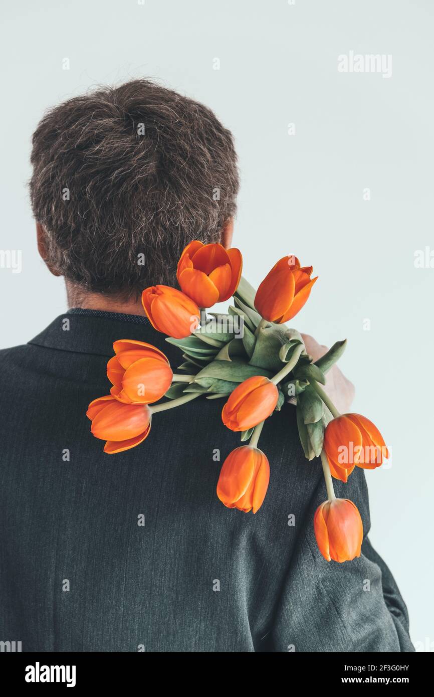 Ein eleganter Mann mittleren Alters trägt einen Strauß oranger Tulpen auf seiner Schulter. Überraschung für Ihre geliebte Frau. Romantisches Kurzurlaub-Konzept. Rückansicht. Stockfoto