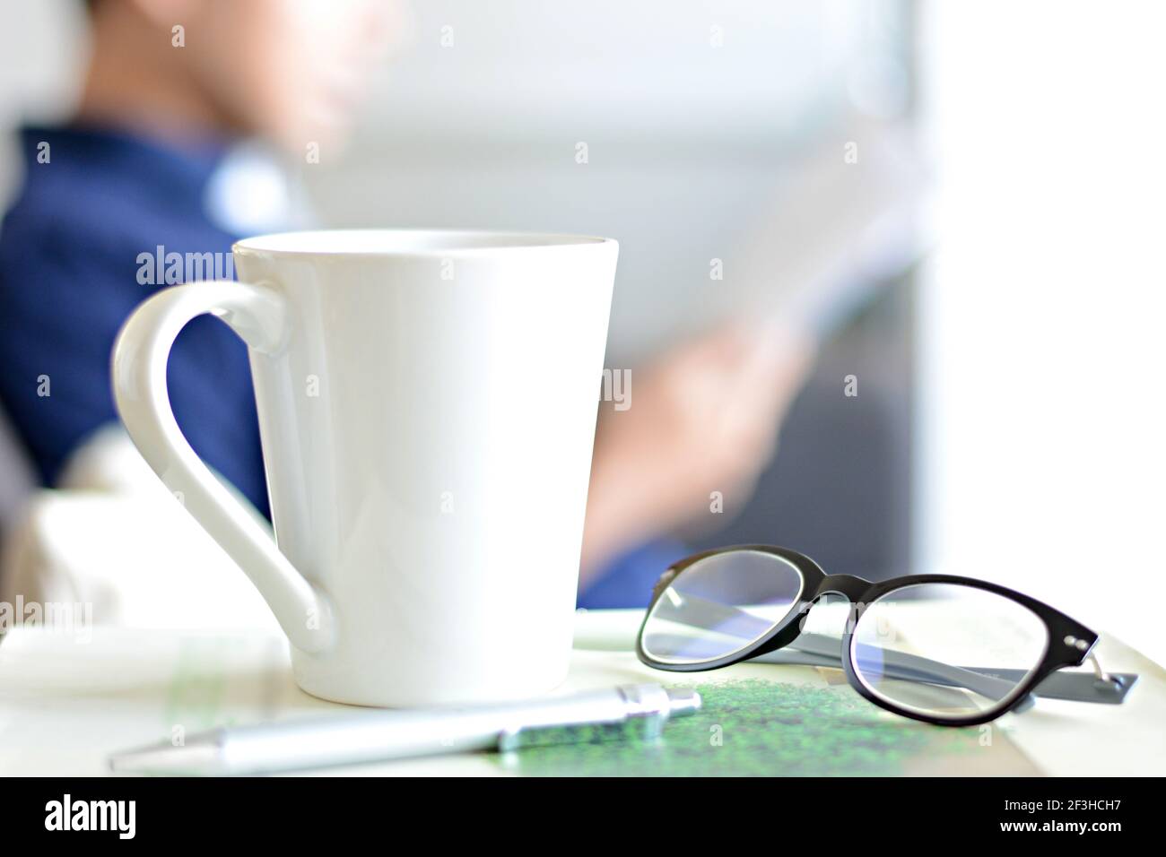 Kaffeetasse, Augenbrille & Stift über einem Buch mit unscharfen Hintergrund eines Mannes Buch zu lesen Stockfoto