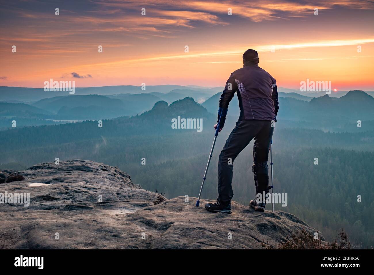 Behinderter Tourist mit Krücken. Rückansicht des verletzten Wanderers mit Unterarmstangen, die vor dem Hintergrund des Sonnenuntergangs am Berg laufen Stockfoto
