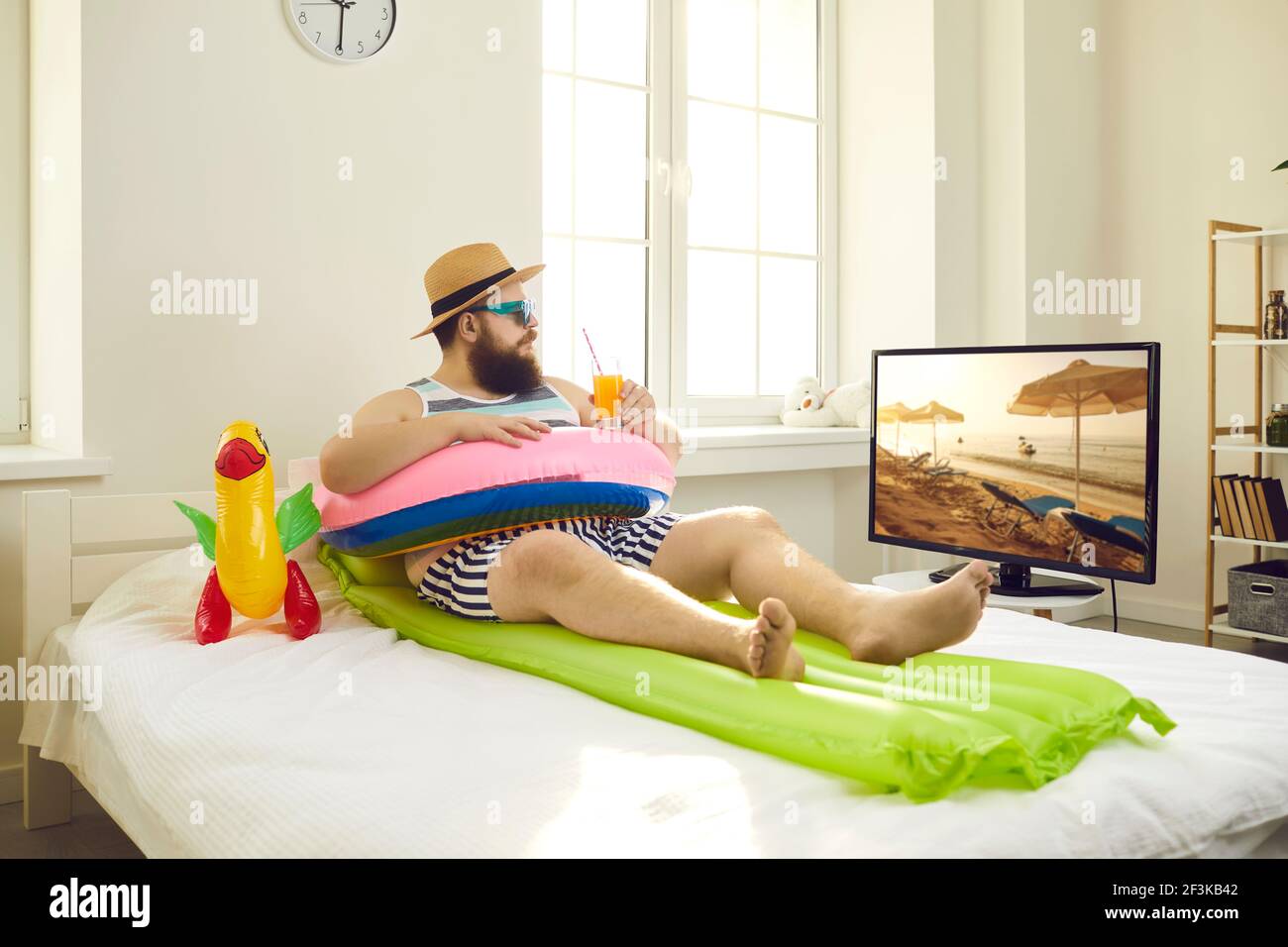 Lustiger Mann sitzt auf dem Bett mit aufblasbarem Schwimmring, nippt Saft und schaut Reise-TV-Show Stockfoto