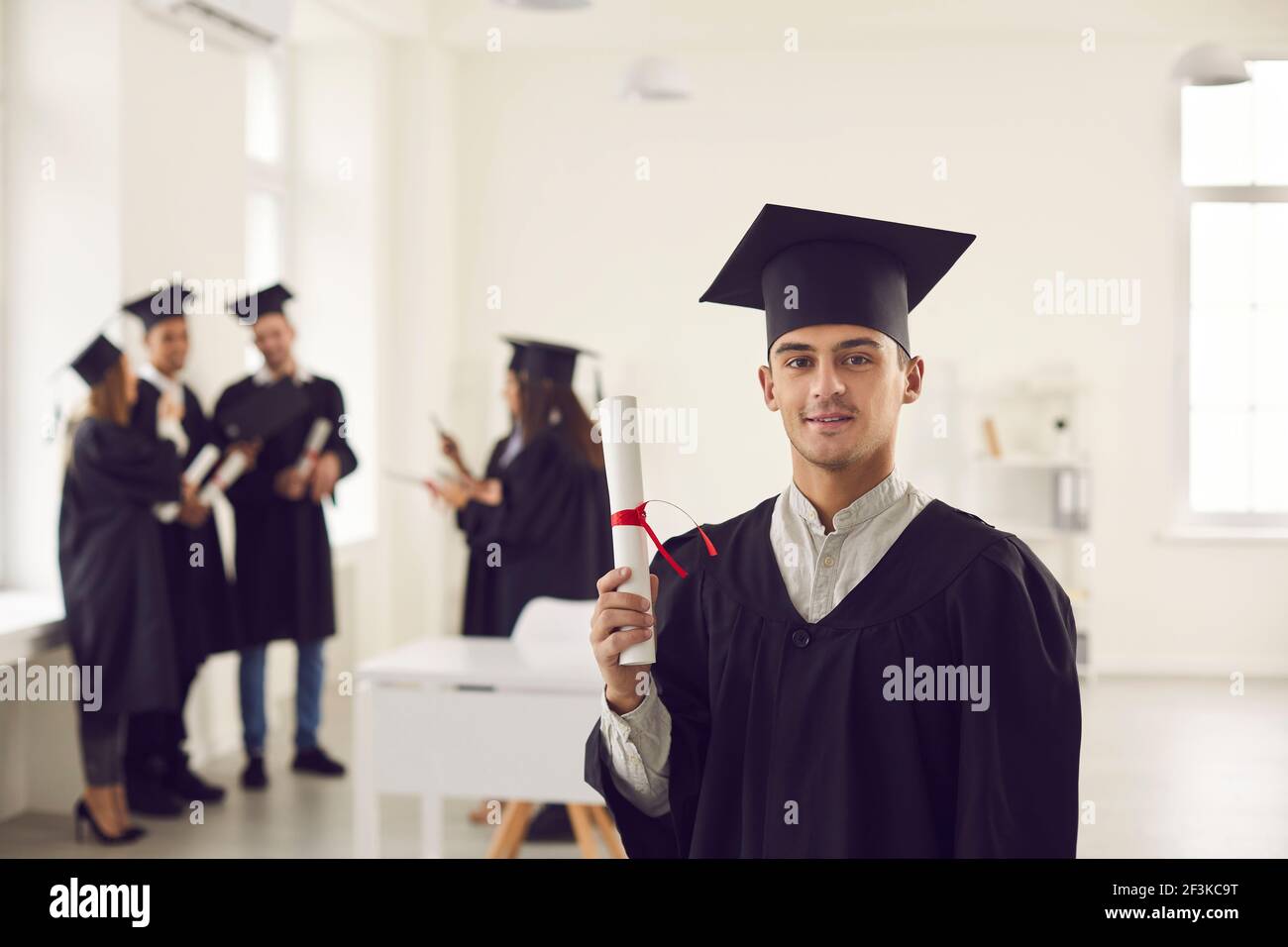 Selbstbewusster Student Kerl in der Uniform eines Hochschulabsolventen zeigt ein Diplom in seinen Händen. Stockfoto