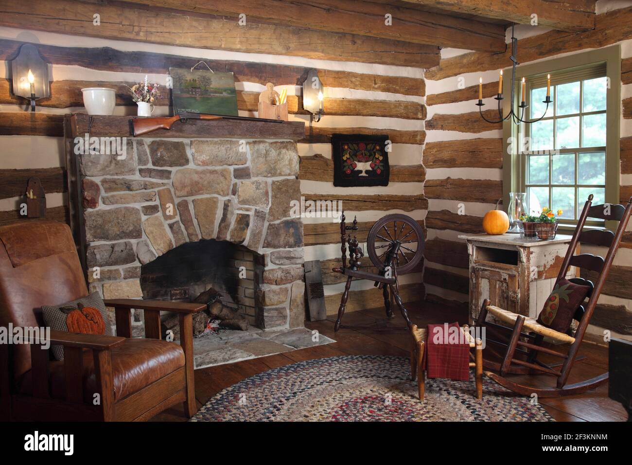 Wohnzimmer in historischer Blockhütte mit Steinkamin und freiliegenden Holzstämmen. Schaukelstuhl, Mission-Stil Stuhl und Spinnrad, zusammen mit Teppich. Stockfoto
