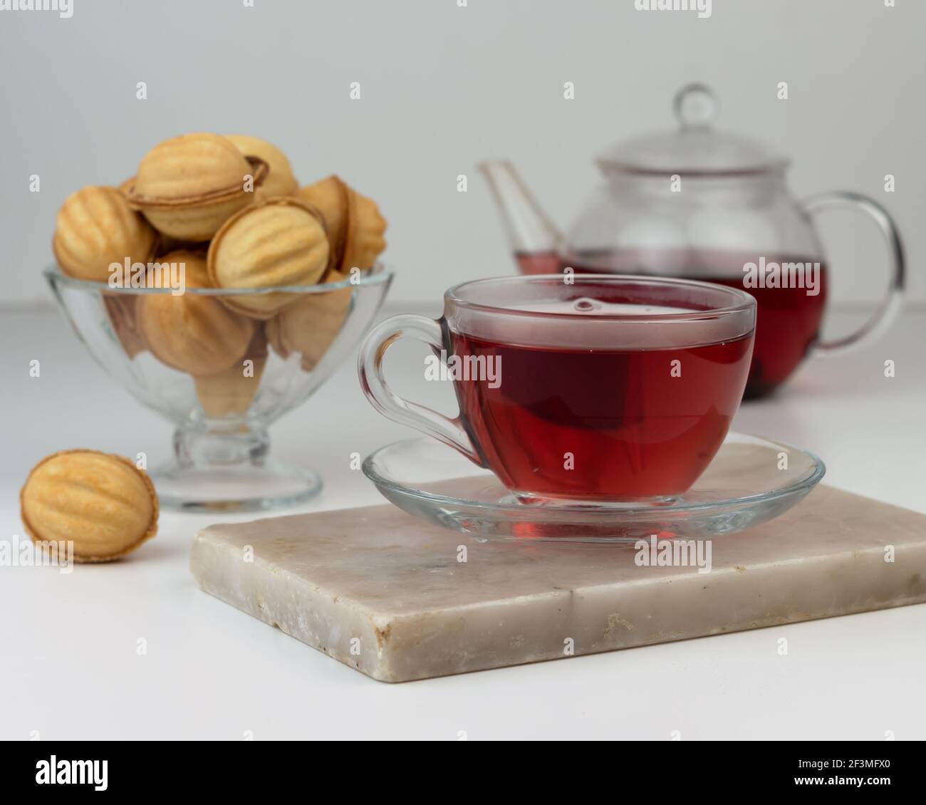 Hausgemachte, nussförmige Kekse, gefüllt mit Kondensmilch in einer Glasvase auf einem Schneidebrett aus Marmor. Roter sudanesischer Rosetee in einer Tasse und Teekanne. Stockfoto