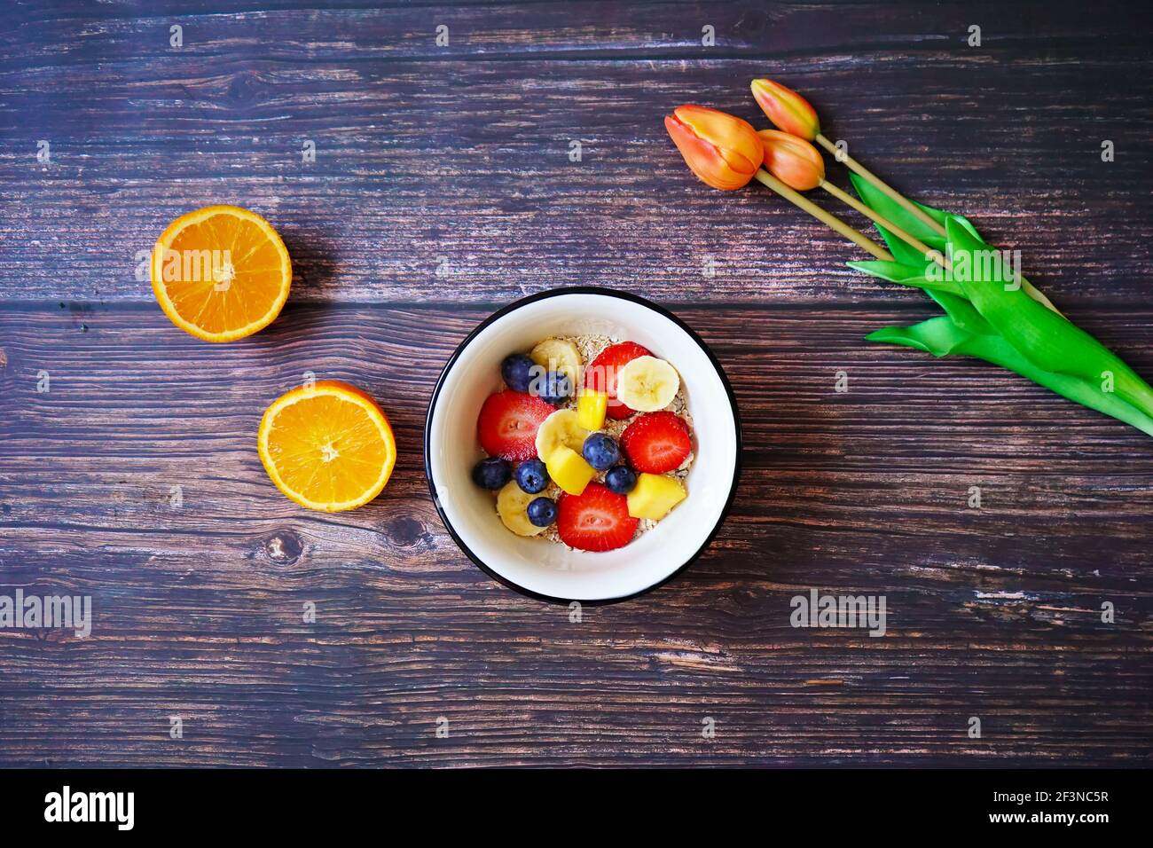 Eine Müslischale mit frischen Früchten (Erdbeere, Banane, Heidelbeere, Mango) und einer Orange auf einem hölzernen Frühstückstisch. Orangefarbene Tulpen zur Dekoration. Stockfoto