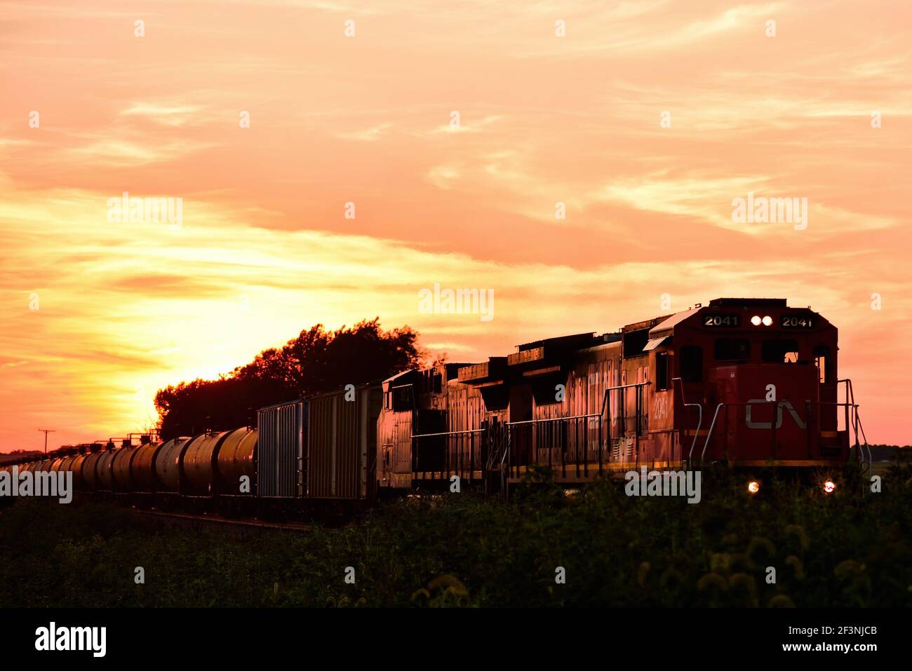 Burlington, Illinois, USA. Hinter dem Sonnenuntergang fährt ein Güterzug, der in Richtung Chicago fährt, durch das ländliche Illinois. Stockfoto