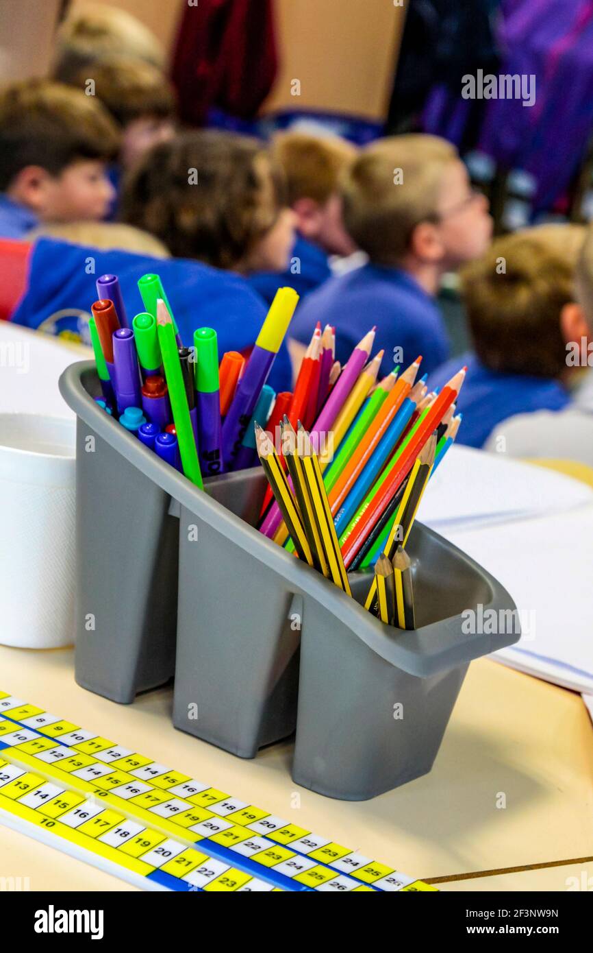 Grauer Kunststoffhalter mit Stiften und Bleistiften mit Grundschulkindern in einem Klassenzimmer im Hintergrund. Stockfoto