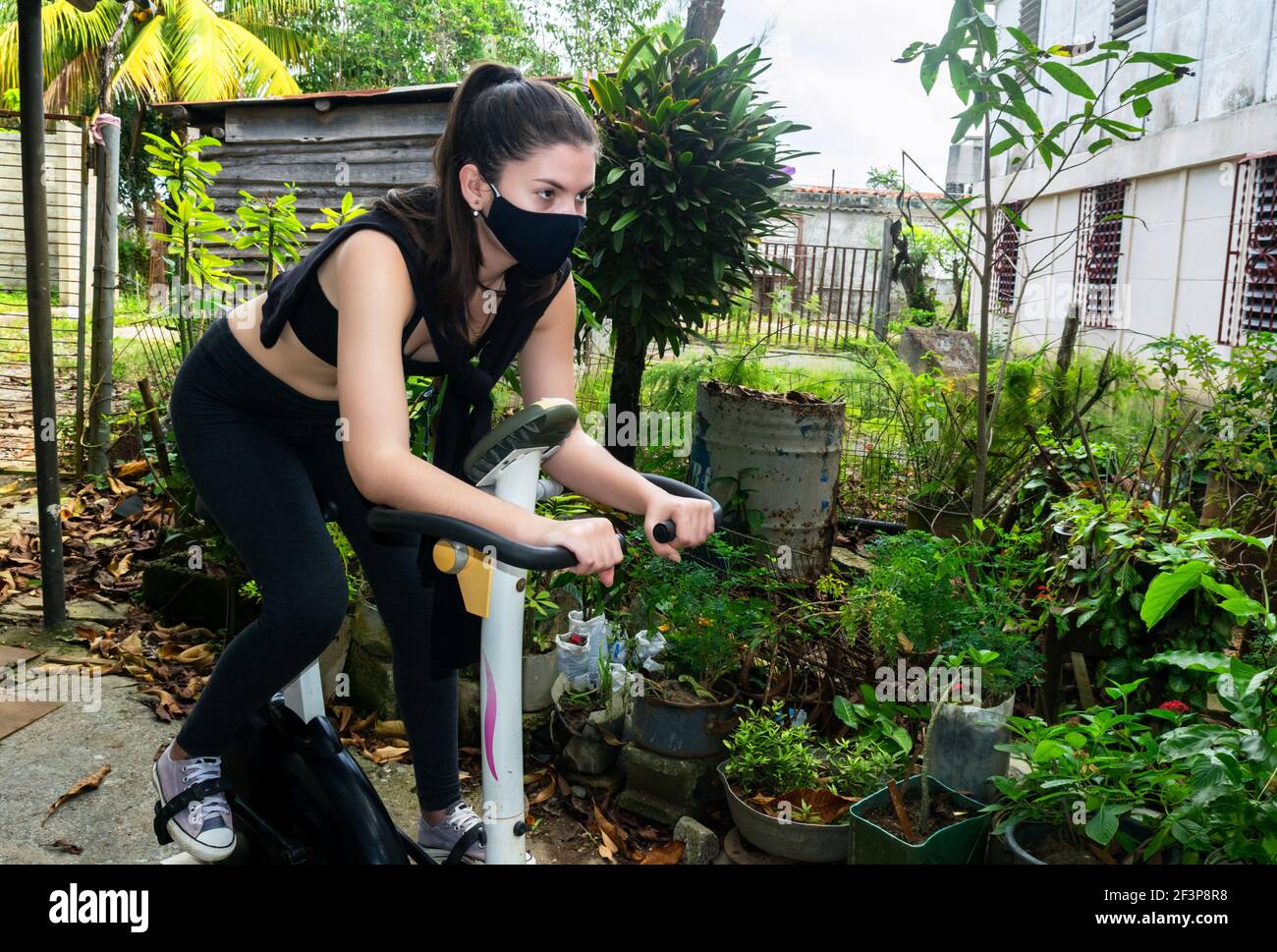 Junge kubanische Frau spinnen auf einem statischen Fahrrad in einem Hinterhof während Pandemiezeiten Stockfoto