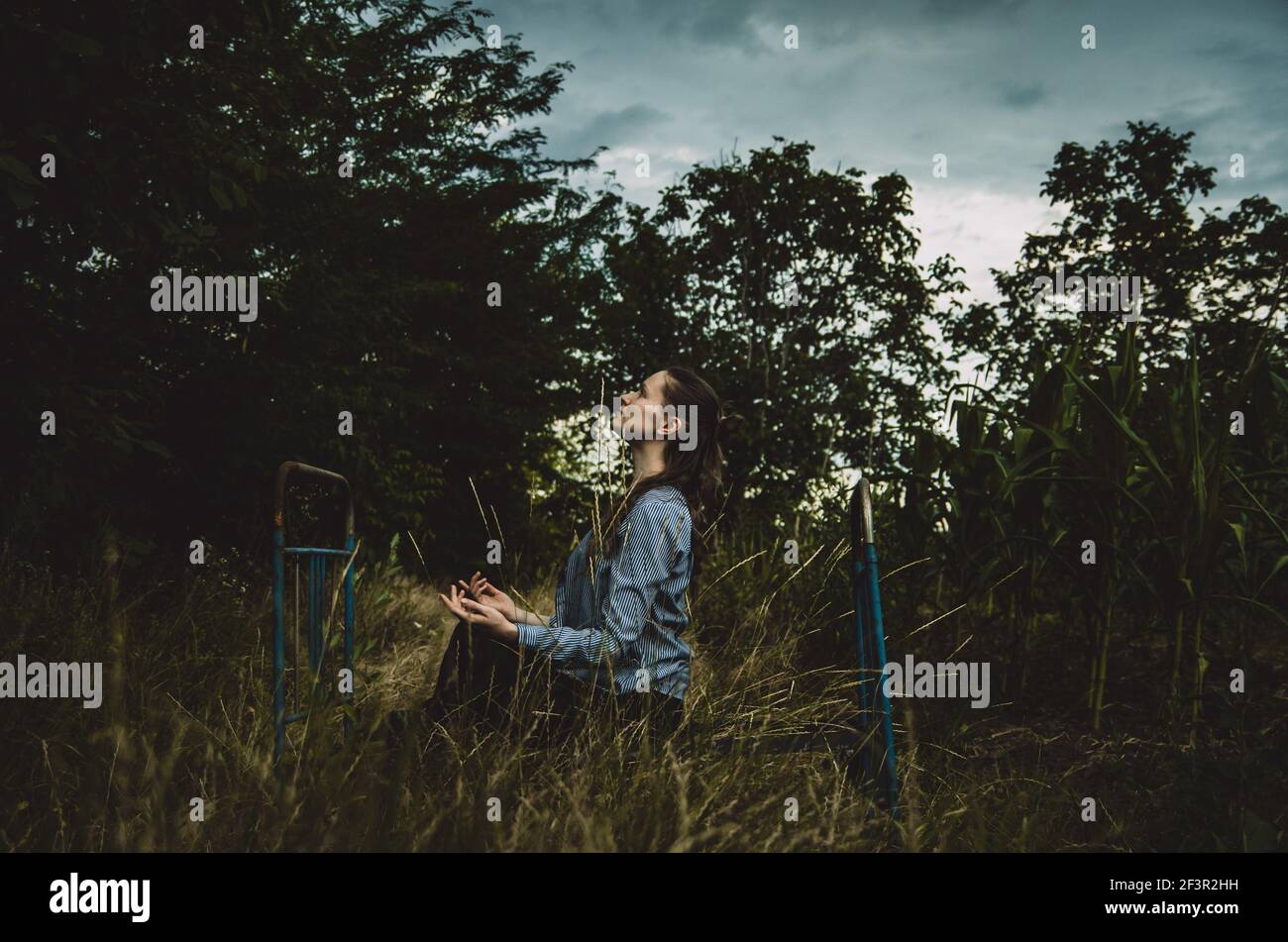 Junge Frau, die auf einem alten Metallbett sitzt und inmitten der Natur meditiert, mit launischer dunkelgrüner Atmosphäre Stockfoto