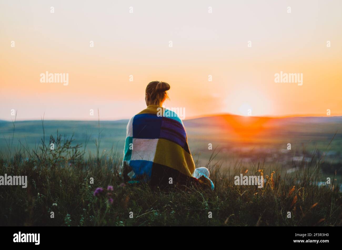 Rückansicht einer jungen Frau, eingewickelt in eine bunte Strickdecke, sitzend auf einem Hügel bei Sonnenuntergang, meditierend, kontemplierend Stockfoto