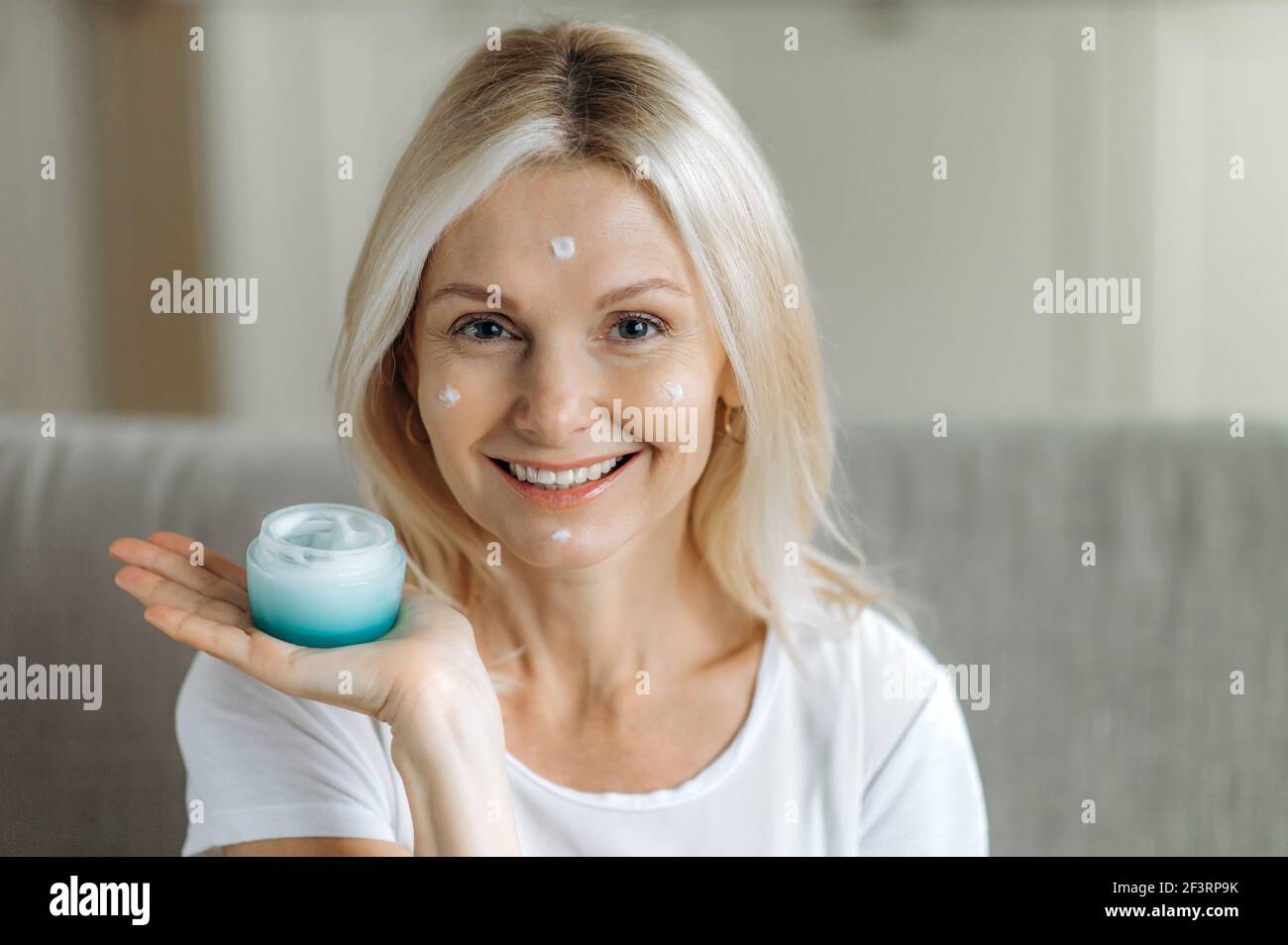 Kaukasische blonde reife Frau lächelt und schaut auf die Kamera. Erstaunliche Erwachsene Dame hält eine Creme in der Hand, feuchtigkeitsspendende Gesichtscreme in ihrem Gesicht, trägt Tropfen der Creme auf ihr Gesicht, Hautpflege Stockfoto