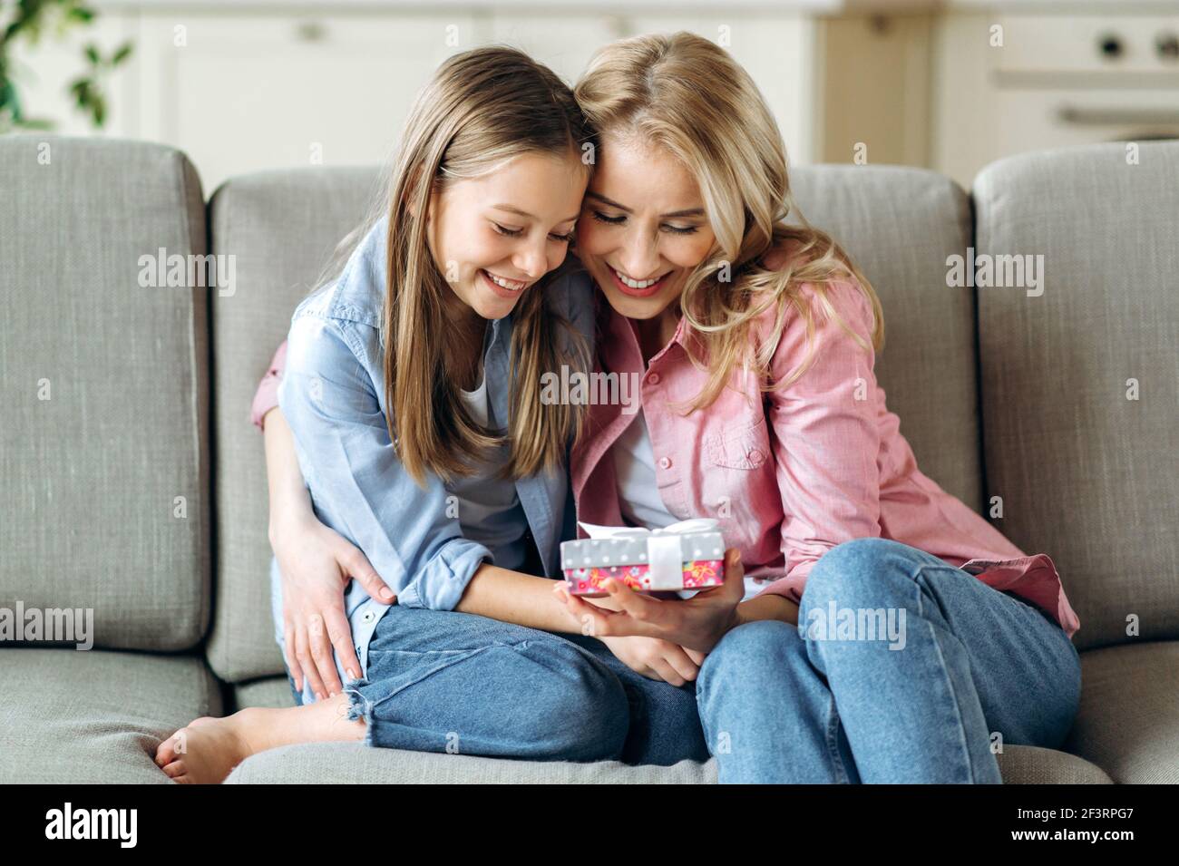 Mutter und Tochter umarmen sich gegenseitig. Eine blonde kaukasische Mutter und ihr Vorschulmädchen sitzen auf dem Sofa im Wohnzimmer und schauen auf die Geschenkbox, wunderschön dekoriert mit einer Schleife, lächelnd Stockfoto