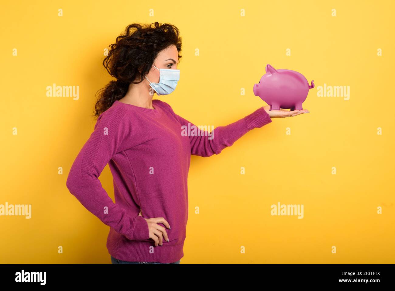 Nachdenkliche Frau mit Gesichtsmaske hält ein Sparschwein. Konzept der Geldeinzahlungsbank. Gelber Hintergrund Stockfoto