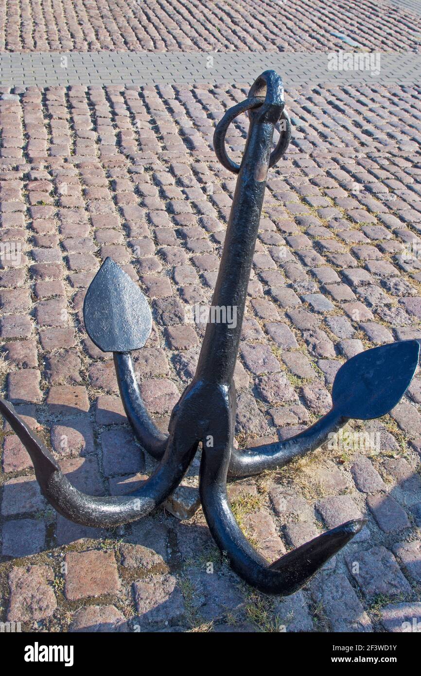 Ein alter vierbeiniger Anker mit mandelförmigen Punkten an Die Enden schwarz lackiert mit einem Ring an der Spitze Stehend auf einem gepflasterten Platz von rauem braunen Pflaster Stockfoto