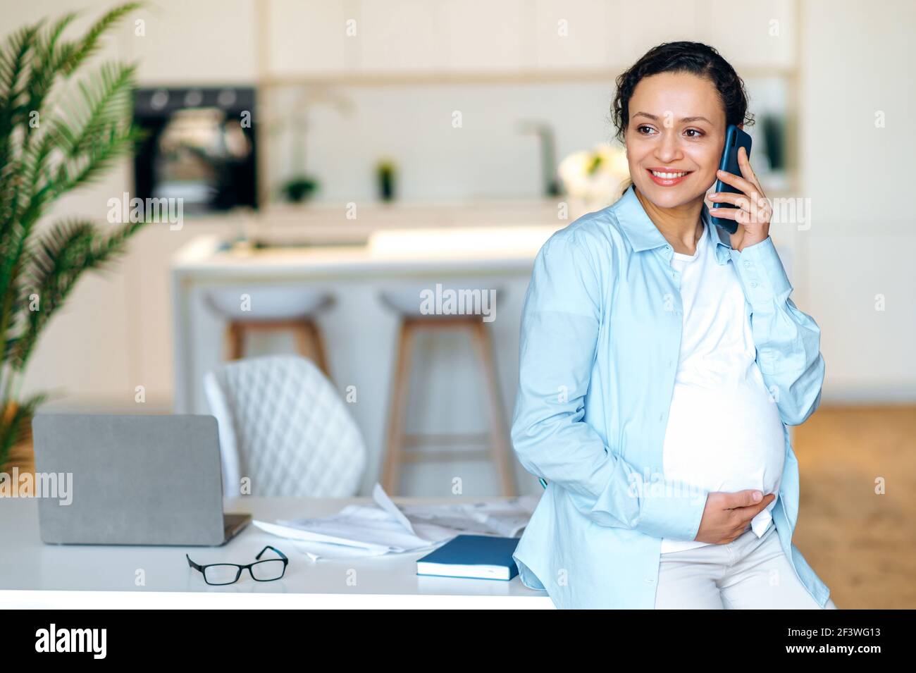 Erfolgreiche attraktive gemischte Rasse schwangere Erwachsene Frau steht in der Nähe ihres Arbeitsplatzes, mit angenehmen Handy-Gespräch, sanft berühren ihren Bauch, wegschauen und lächeln Stockfoto