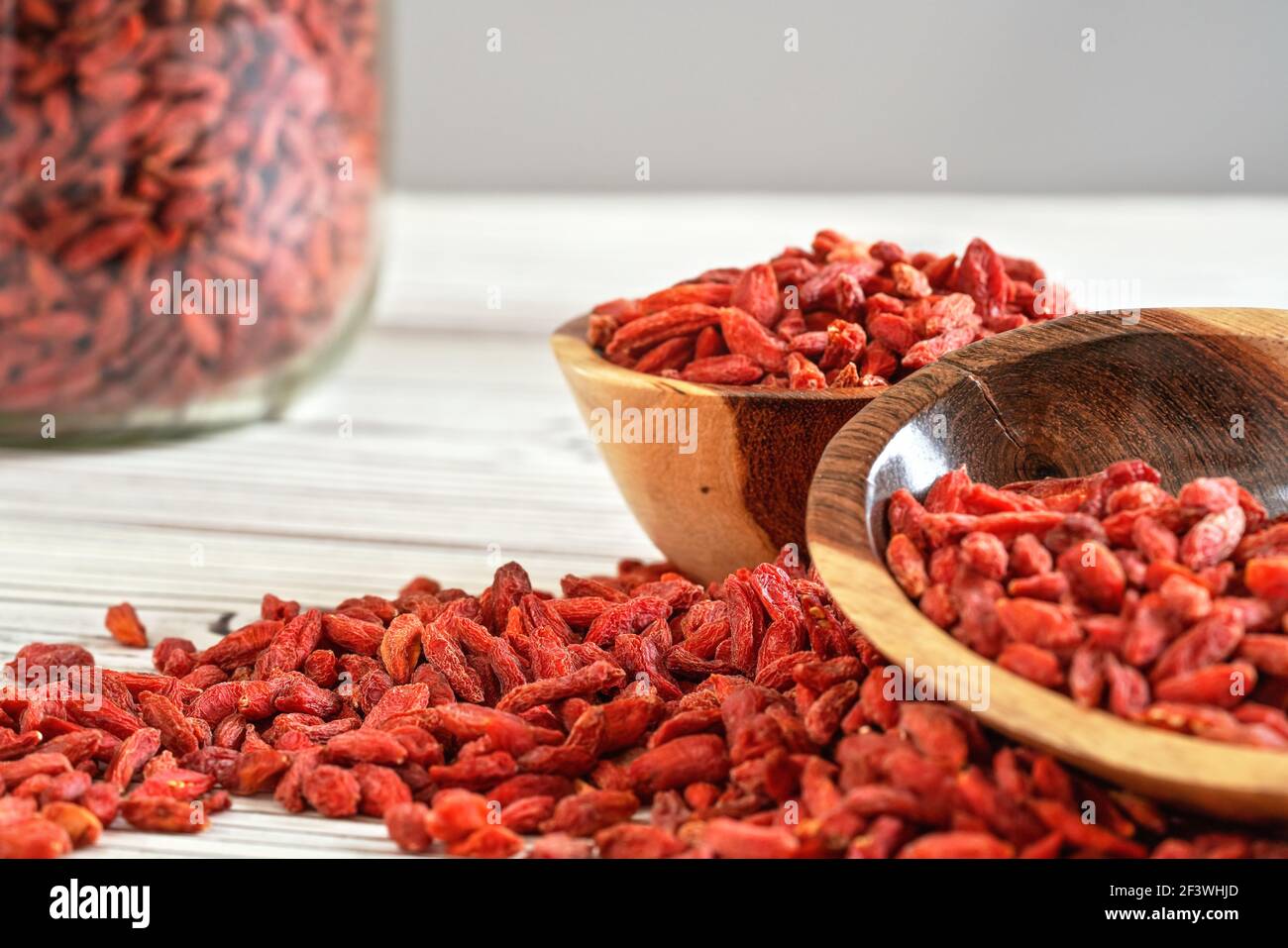 Getrocknete Goji Beeren in Holzschalen, verstreut über weißen Brettern Tisch unter, verschwommen großen Glas voller Früchte im Hintergrund Stockfoto