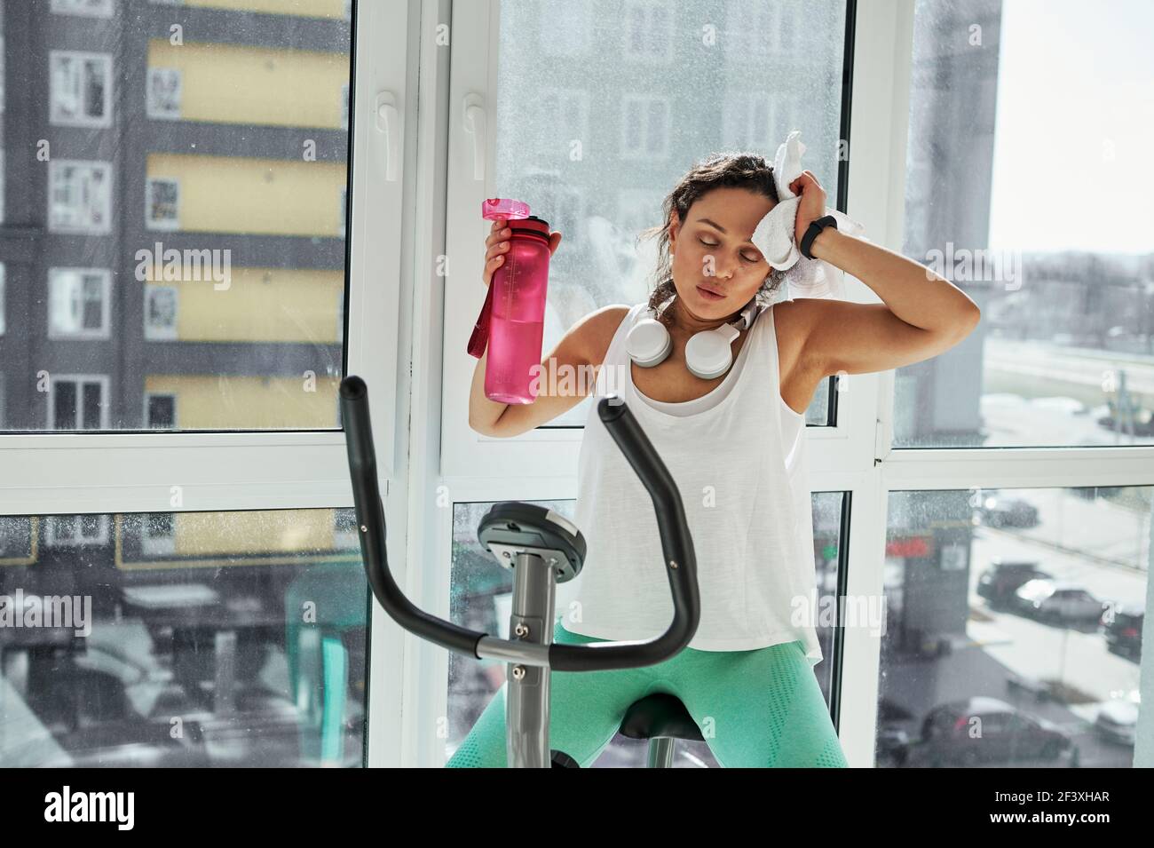 Eine junge Frau mit gemischtem Rennen wischt ihr Gesicht mit einem Handtuch, erschöpft von einem schweren Cardio-Training. Zeit zum Ausruhen nach dem Ergometer Stockfoto