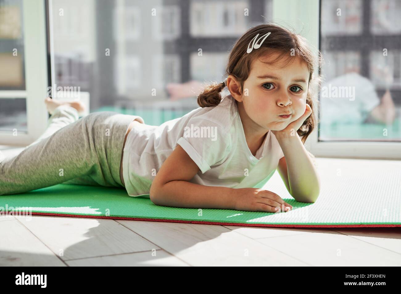 Liebenswert kleines Mädchen auf einer Übungsmatte entspannen und posieren Vor dem Hintergrund großer Fenster zu Hause fotografieren An einem sonnigen Tag Stockfoto