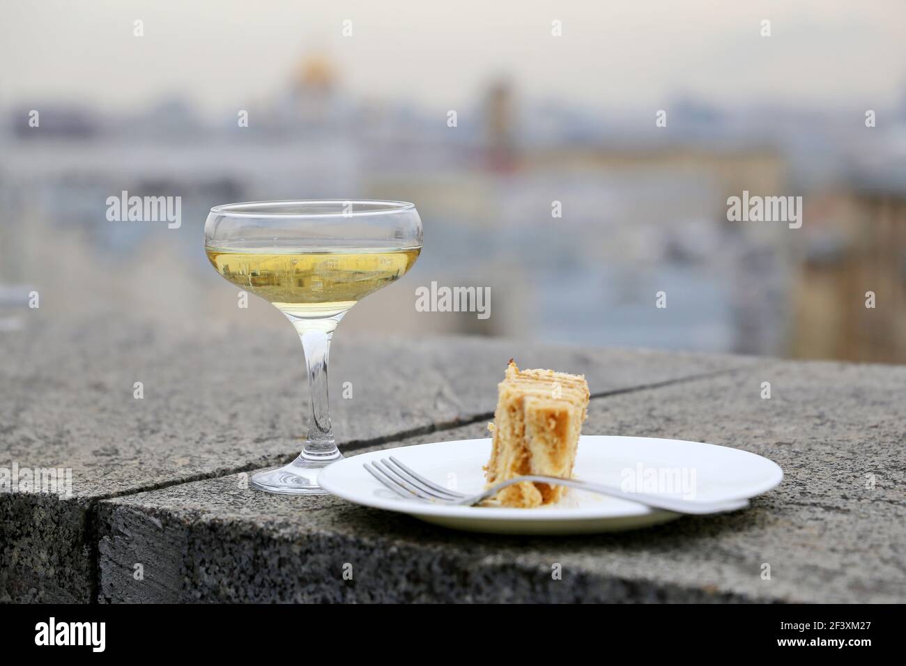Glas Sekt und Teller mit süßem Kuchen auf der Steinbrüste im Stadthintergrund. Konzept der Reise, Feier, romantisches Abendessen Stockfoto