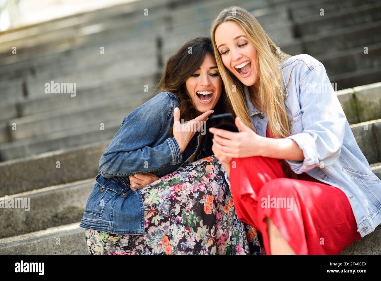 Zwei junge Frauen an etwas lustige Sache auf Ihre Smart Phone draussen, sitzen auf den städtischen Schritte. Freunde Mädchen lachend. Stockfoto